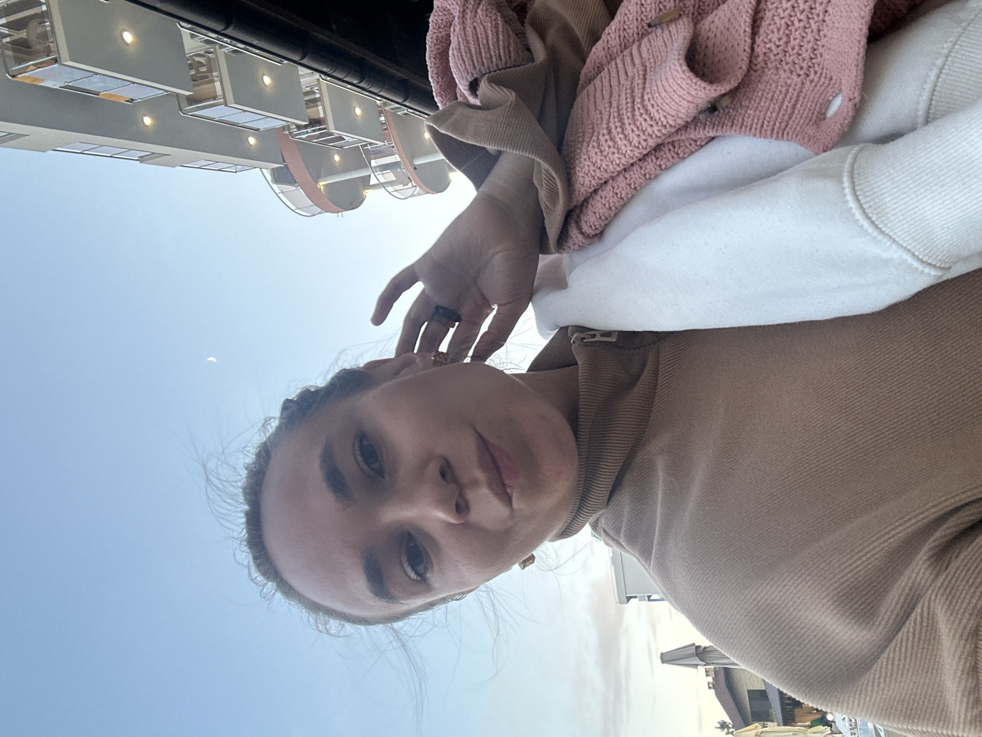 A young woman with brown hair pulled back, wearing a tan turtleneck sweater and earrings, is taking a selfie outdoors. She is holding a pink knitted cardigan and a white sweatshirt in her arms. Her hand is raised to her ear, and she is wearing a ring. In the background, a modern building with balconies and lights is visible, along with a crescent moon in a clear sky. The scene appears to be set in Vityazevo, Russia, with hints of urban architecture.