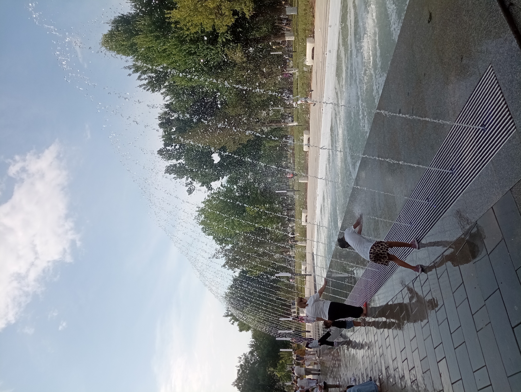In Shymkent, Kazakhstan, a group of people are enjoying a sunny day at a modern water fountain. The scene is vibrant with the clear blue sky and lush green trees in the background. The fountain consists of many jets of water that create a shimmering curtain across the paved area. Several individuals, including children and adults, are actively interacting with the water, some walking through it while others are captured mid-splash.
One person, wearing a white t-shirt and patterned shorts, is seen lying on a striped metal grate, letting the water spray over them. Another person in a white t-shirt and black pants is also near the water jets, with their hands on their knees. Further back, more people are visible, enjoying the cooling spray. The ground around the fountain is wet, reflecting the light and the surrounding trees. The time of day appears to be afternoon, given the bright sunlight and clear sky, suggesting pleasant weather for outdoor recreation. The overall mood seems joyful and relaxed, with people of all ages participating in the fun. There is no visible text in the image.