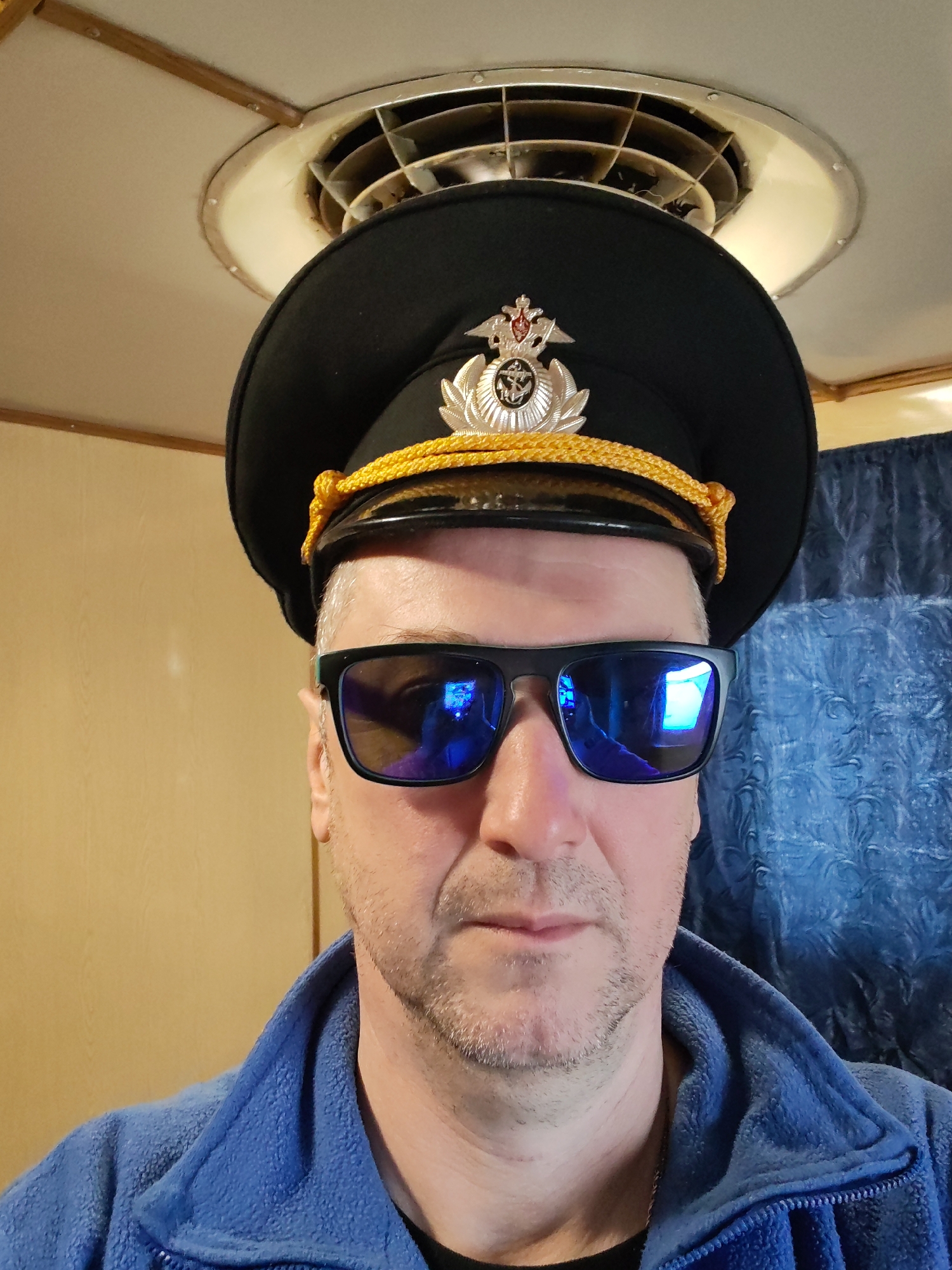 A close-up indoor portrait features a middle-aged man wearing a black uniform cap with a gold braid and a prominent metallic badge, likely a Russian naval or maritime insignia featuring a double-headed eagle and anchor. He is also wearing black-framed sunglasses with blue reflective lenses and a blue fleece jacket over a dark shirt. The man has short, light-colored hair and stubble on his face.
The background shows a light-colored ceiling with a circular vent or light fixture directly above his head, featuring a spiral-like design. Wooden trim is visible along the ceiling line. To the man's right, a textured blue curtain or fabric is present, while a plain light brown wall is to his left. The reflections in his sunglasses suggest internal light sources. The setting appears to be an interior space, possibly on a boat or within a facility. The activity is a posed photograph. No specific emotions are discernible due to the sunglasses. The location context provided is Bol'shie Mkhi, Russia. No visible text is present in the image.
