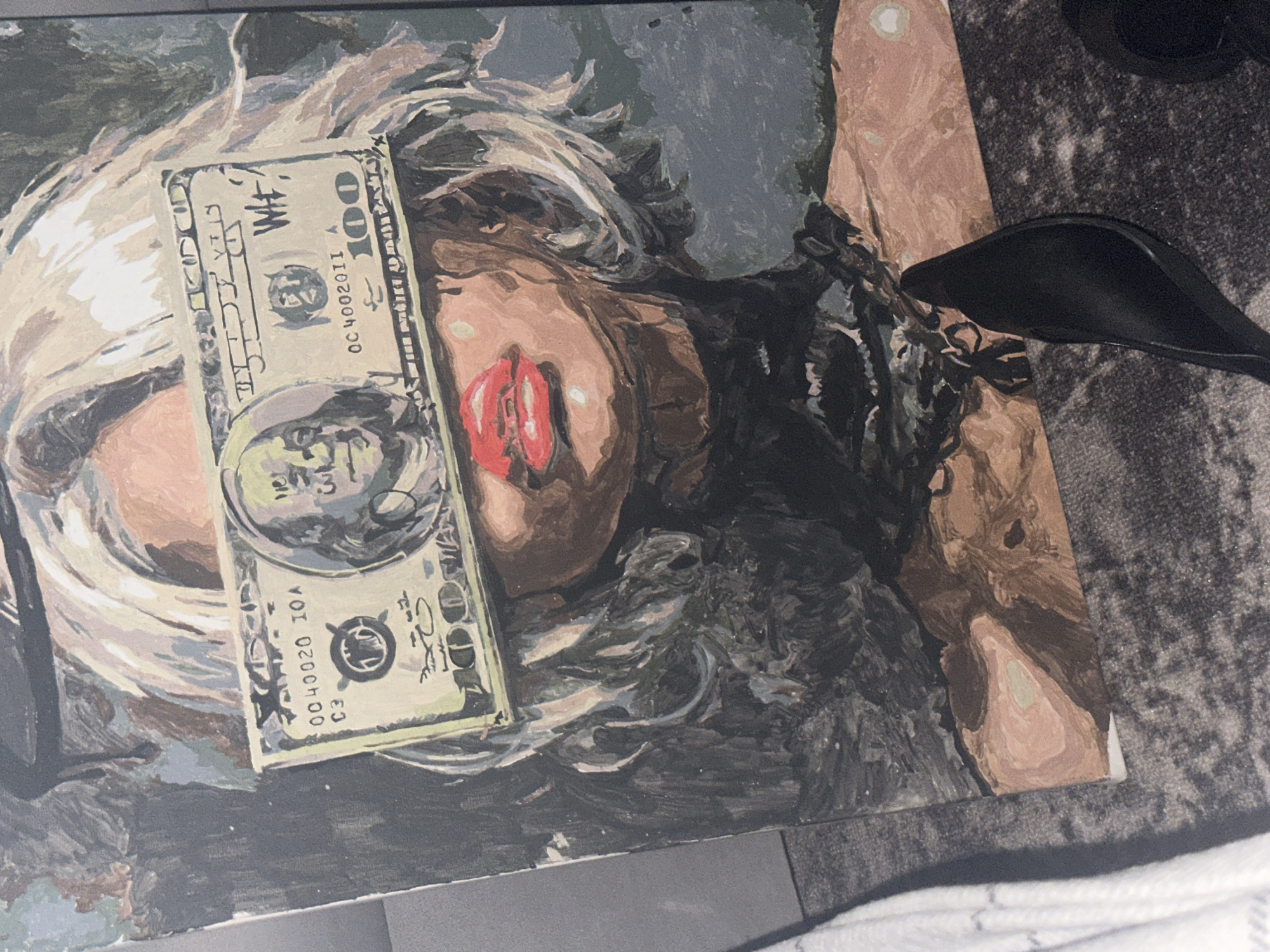 A painting depicts a person with blonde hair and red lips, with a stylized $100 bill covering part of their face. The person is wearing what appears to be a black lace garment. The painting is lying on a surface, partially on a gray, patterned rug. A black shoe is visible on the rug. The location is Высокий, Russia.