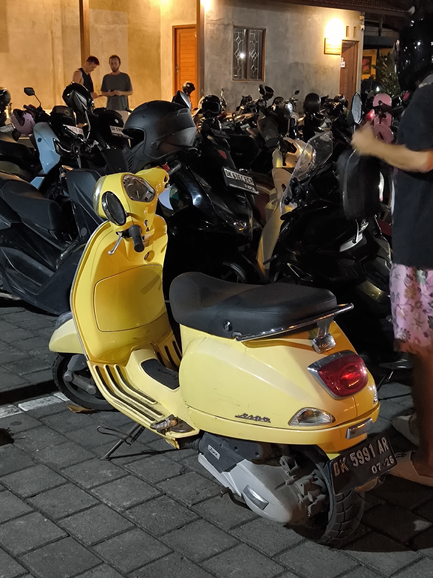 A yellow Vespa scooter is parked amongst a dense row of other motorcycles in Canggu, Indonesia. Two men stand near the front of the rows of motorcycles, observing something, with a building behind them. The Vespa has a license plate that reads "DK 5991 AR" and a date sticker of "07-26". A person wearing floral-patterned shorts and white shoes stands adjacent to the Vespa's rear. The area is paved with interlocking gray bricks, and the scene is illuminated with artificial lighting.