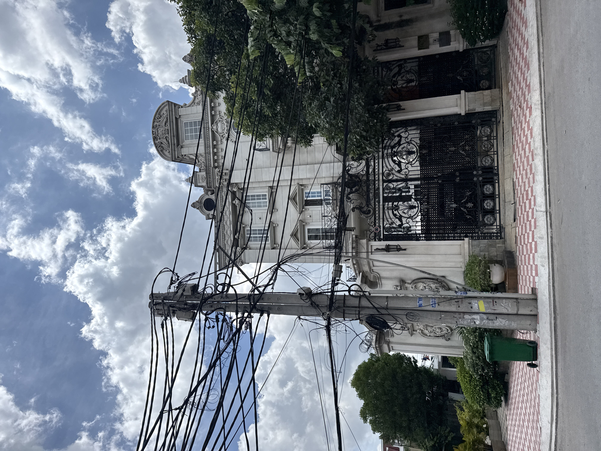 In Thủ Đức City, Vietnam, a stately white building with ornate architectural details stands behind a wrought-iron gate. The building features multiple windows, balconies with railings, and decorative elements on its facade. A large, elaborate gate with intricate scrollwork and floral motifs guards the entrance. Green trees and manicured bushes surround the property, providing a lush contrast to the white structure. In the foreground, a gray utility pole is heavily encumbered with a dense network of black electrical wires, creating a stark visual juxtaposition with the elegant building. A green trash bin is positioned near the base of the utility pole, and the ground is paved with red and white checkered tiles. The sky above is partly cloudy, with fluffy white clouds against a blue backdrop.