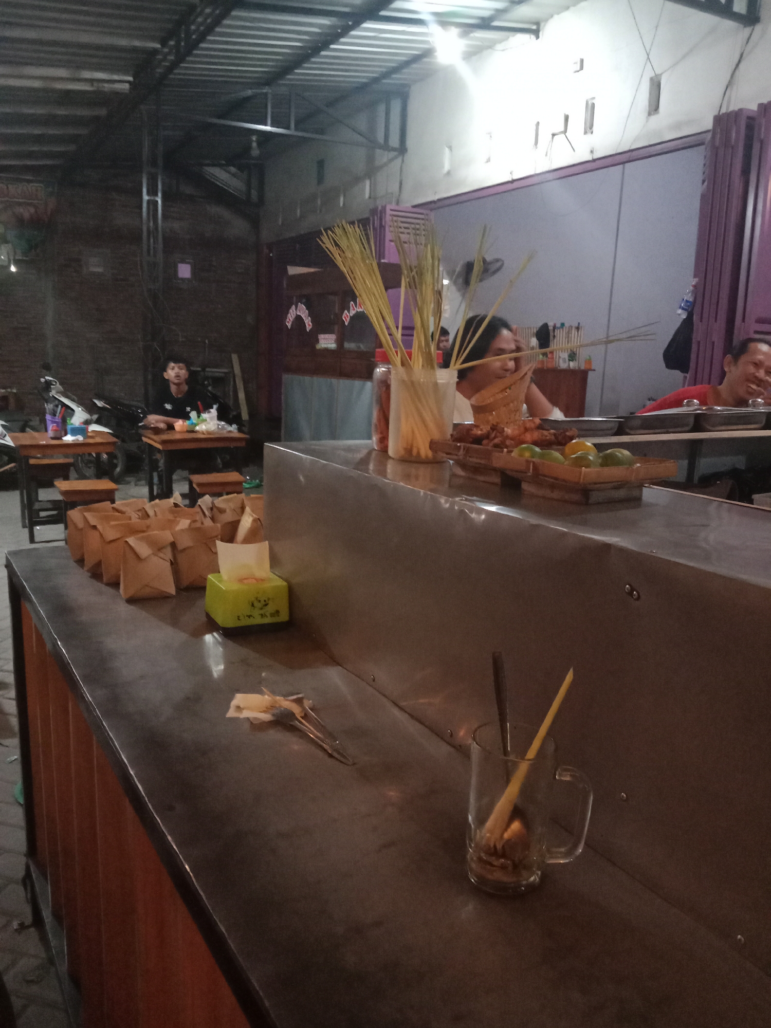 This image depicts a casual dining establishment, likely a warung or small restaurant, located in Cangkring, Indonesia, captured in low-light conditions. The scene primarily focuses on a service counter in the foreground and patrons seated at tables in the midground and background. In the foreground, a metallic counter extends diagonally from the lower-left to the right. On the left side of the counter, a stack of approximately twelve small, brown paper bags, folded and sealed, are neatly arranged. Adjacent to these bags is a lime green tissue box with white text. Further right, a clear glass jar contains numerous thin, light-colored skewers. Behind the jar, parts of a clear plastic bottle and a red condiment bottle are visible. A wooden tray containing several pieces of grilled food, possibly chicken or other meat, and four green limes is positioned on the counter, slightly to the right of the skewers. In the bottom right corner of the counter, a clear glass mug with a handle holds a brown spoon and a yellow stirrer. Near the center of the counter, a crumpled piece of paper or tissue lies beside a pair of small metal tongs. In the midground, three individuals are present. To the left, a young male with short dark hair, wearing a dark t-shirt, is seated at a simple wooden table with a wooden stool. He is looking upwards and slightly to the right. A small white container and other indistinct items are on his table. Behind him, a motorbike is partially visible. Centrally, behind the service counter, a person with long dark hair is seated, looking down with a slight smile, seemingly engaged with the items on the counter. To the right, another male with short dark hair, wearing a red t-shirt, is seated at a different wooden table, laughing with an open mouth and looking towards the right side of the frame. The background reveals the establishment's structure. On the left, an exposed brick wall is visible, with a metal support beam extending vertically. The ceiling features a corrugated metal roof with visible metal trusses. On the right side of the background, a light grey wall with several small, vertical rectangular cutouts is present. Further right, a purple, possibly corrugated metal or plastic, structure resembling a gate or partition is visible. Above this, red and white triangular banners are strung across. The overall lighting is dim, suggesting evening hours,