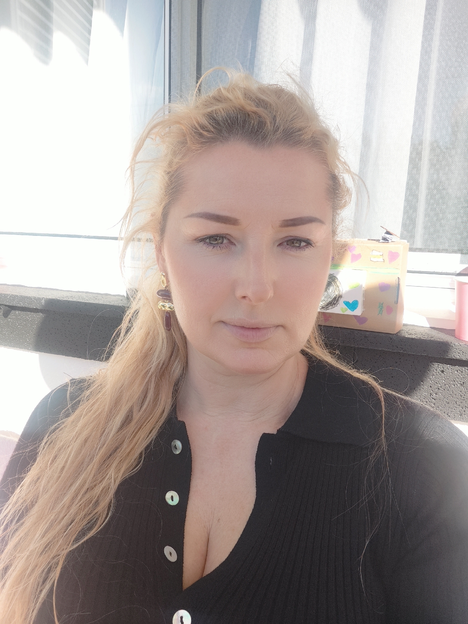 A woman with fair skin, light blonde hair pulled back loosely, and wearing a black cardigan with pearl buttons is in an interior location in Ribeira Grande, Portugal. She has brown eyes and wears drop earrings with dark red and gold accents. Behind her, on what appears to be a window ledge, there is a decorated cardboard box with drawings of hearts and other designs. There is bright daylight coming through the window and a radiator below the windowsill.