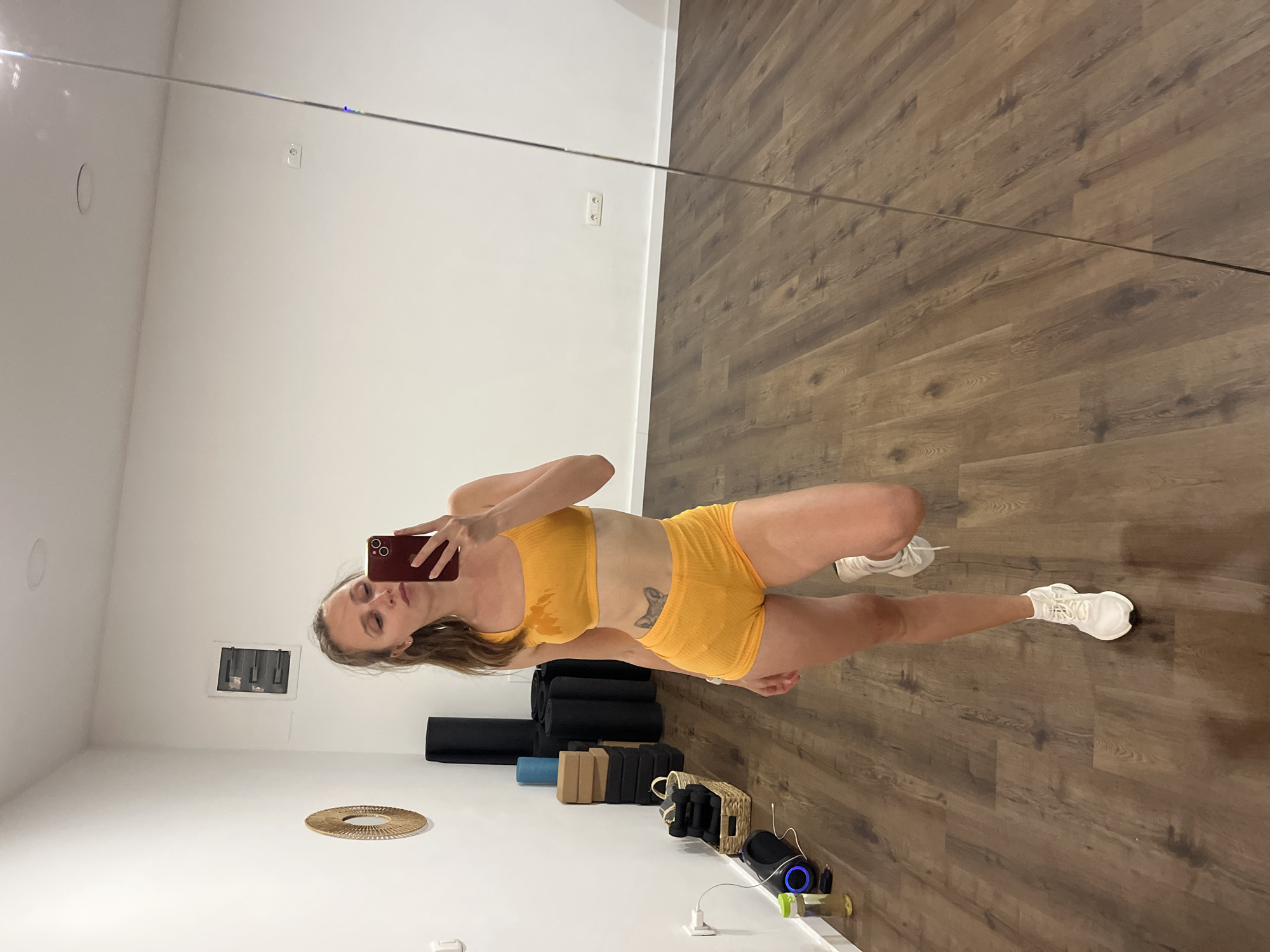 A woman is depicted taking a mirror selfie in what appears to be a fitness studio or home gym. She stands upright, dressed in a bright orange ribbed sports bra and matching high-waisted shorts, paired with white athletic sneakers. Visible sweat marks are present on her sports bra, indicating recent physical exertion. She holds a red smartphone up to capture her reflection, with her head slightly tilted and a neutral expression looking towards the phone. A small, dark tattoo, resembling a dog's head, is visible on her left side abdomen.
The setting features a bright white wall and a warm-toned wooden floor. In the background, various fitness accessories are neatly arranged. Several black yoga mats are rolled up and stacked against the wall, alongside tan and black yoga blocks. A flat, woven, circular mat or decorative item rests on the floor nearby. To the right, a woven basket contains small dumbbells or other exercise equipment. A black speaker with a blue illuminated ring, along with charging cables and adapters, is also visible on the floor. An electrical outlet is present on the white wall. The room is well-lit, suggesting daytime. The image itself is rotated, giving the impression that the woman is lying down, but she is standing in the reflection.