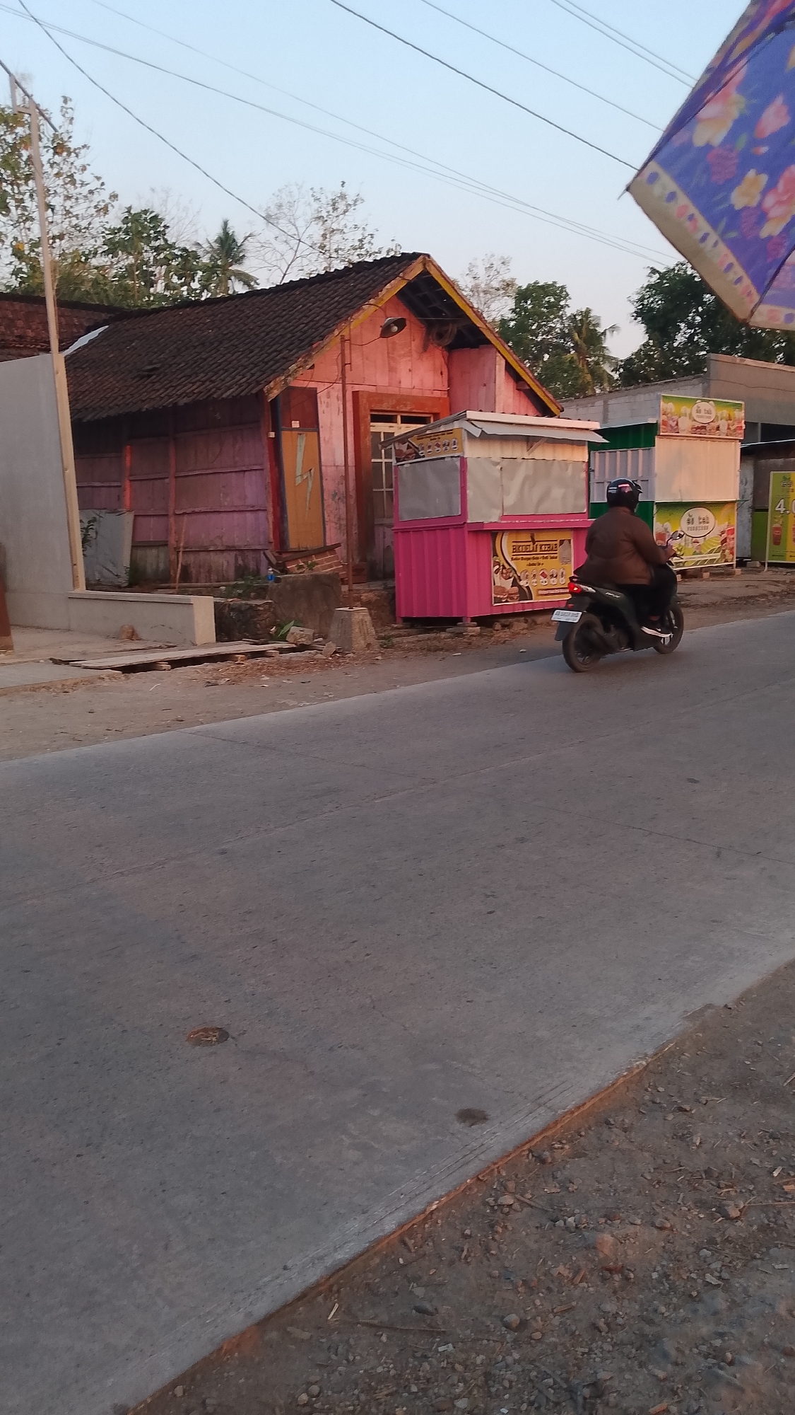 In Karanglangu, Indonesia, a person rides a black motorcycle on a paved road. The motorcycle has a license plate. The rider is wearing a dark jacket and a helmet. In the background, a pink food stall with a white counter is visible, displaying various food items and advertisements. Next to it, another stall is partially visible with a white facade. Behind the stalls, a pink building with a dark roof stands, and further back, trees and clear sky can be seen. A portion of a colorful umbrella or banner is in the upper right corner of the image. The scene appears to be an outdoor market or roadside vendor area.