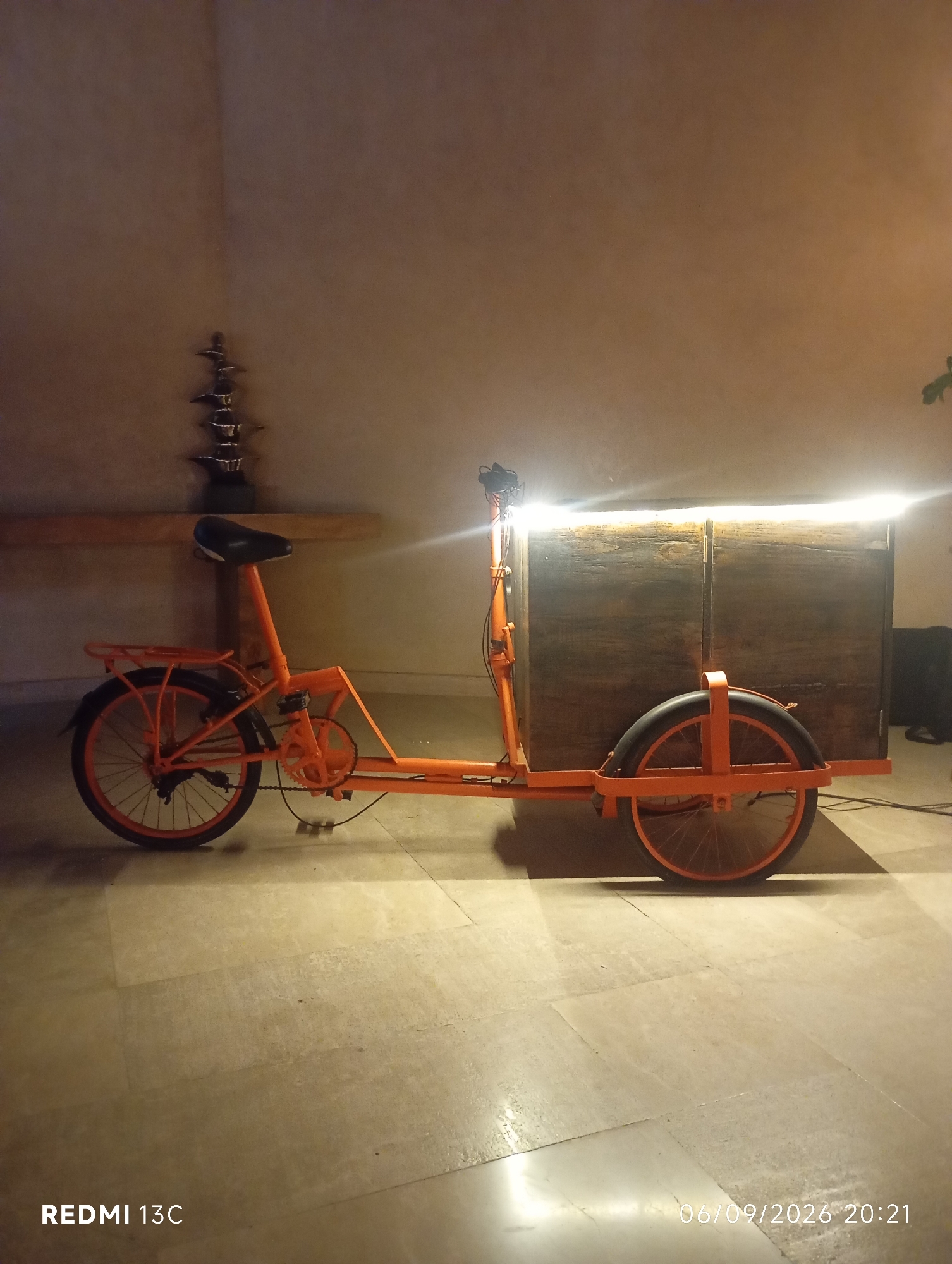 A three-wheeled orange cargo bicycle is parked indoors on a tiled floor, with a wooden box attached to the rear. The bicycle has a black seat, a chain and sprocket system, and fenders on the rear wheels. The wooden box is illuminated from within by a bright light, casting a glow onto the surrounding wall and floor. The wall behind the bicycle is a muted, textured brown. To the left of the bicycle, a multi-tiered decorative object is visible on a wooden shelf. The lighting in the scene is dim, with the primary light source coming from inside the wooden box. The overall impression is one of an indoor setting, possibly a shop or a display area, in Kuta Selatan, Indonesia, given the context of the prompt.