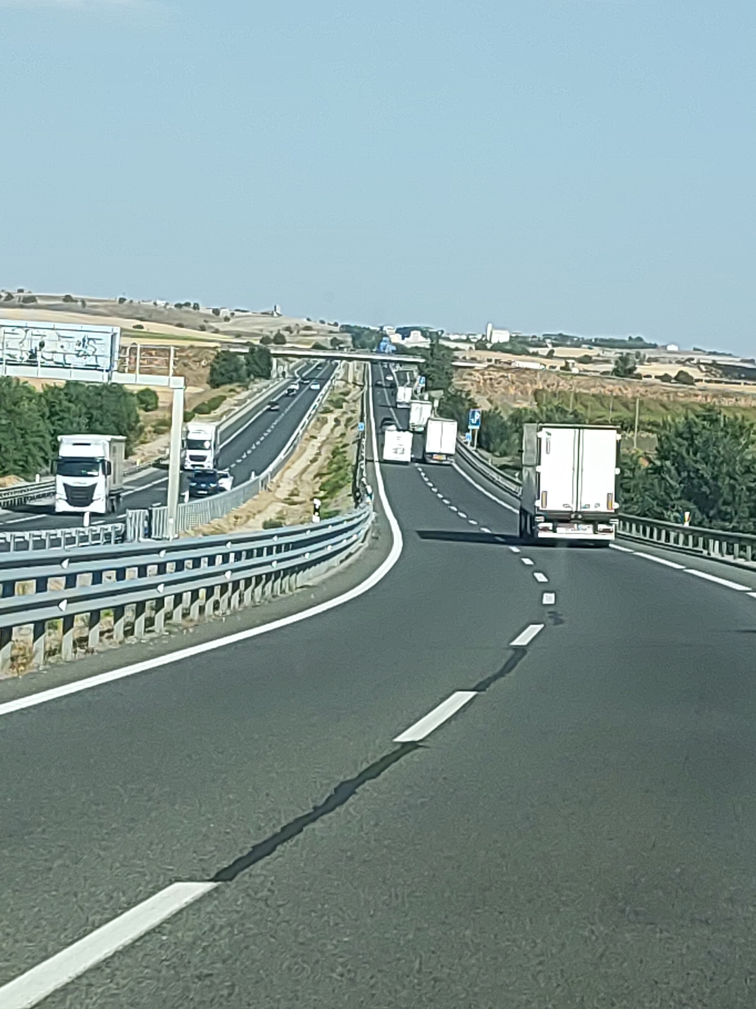 The image shows a highway scene in the city of Almendros, Spain. Several trucks and cars are visible on the road, suggesting moderate traffic. The environment appears to be rural, with dry vegetation and rolling hills in the background. The sky is clear and blue, indicating fair weather. The highway is paved and has white lane markings. A guardrail is present along the side of the road. The perspective suggests the photo was taken from another vehicle traveling on the highway.