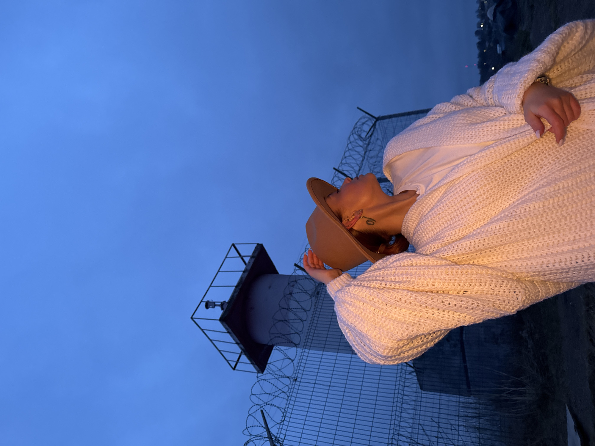 A woman is captured in a full-body, vertically oriented portrait, looking upwards with her head tilted back. She wears a cream-colored, oversized, chunky knit cardigan over a white t-shirt, and a light brown fedora-style hat. A small tattoo is visible on her neck/ear area. Her expression appears pensive or observant as she gazes towards a tall, cylindrical structure in the background.
The setting is outdoors during dusk or night, characterized by a deep, clear blue sky and dim, ambient lighting. In the background, a tall, grey cylindrical tower with a square platform at its top dominates the left side of the frame, possibly a lighthouse or a communication mast. A robust chain-link fence, topped with razor wire, stretches across the middle ground, indicating a secured or industrial area. The overall mood is quiet and contemplative, emphasized by the low light and the woman's upward gaze. There is no visible text in the image. The original image is rotated 90 degrees clockwise.