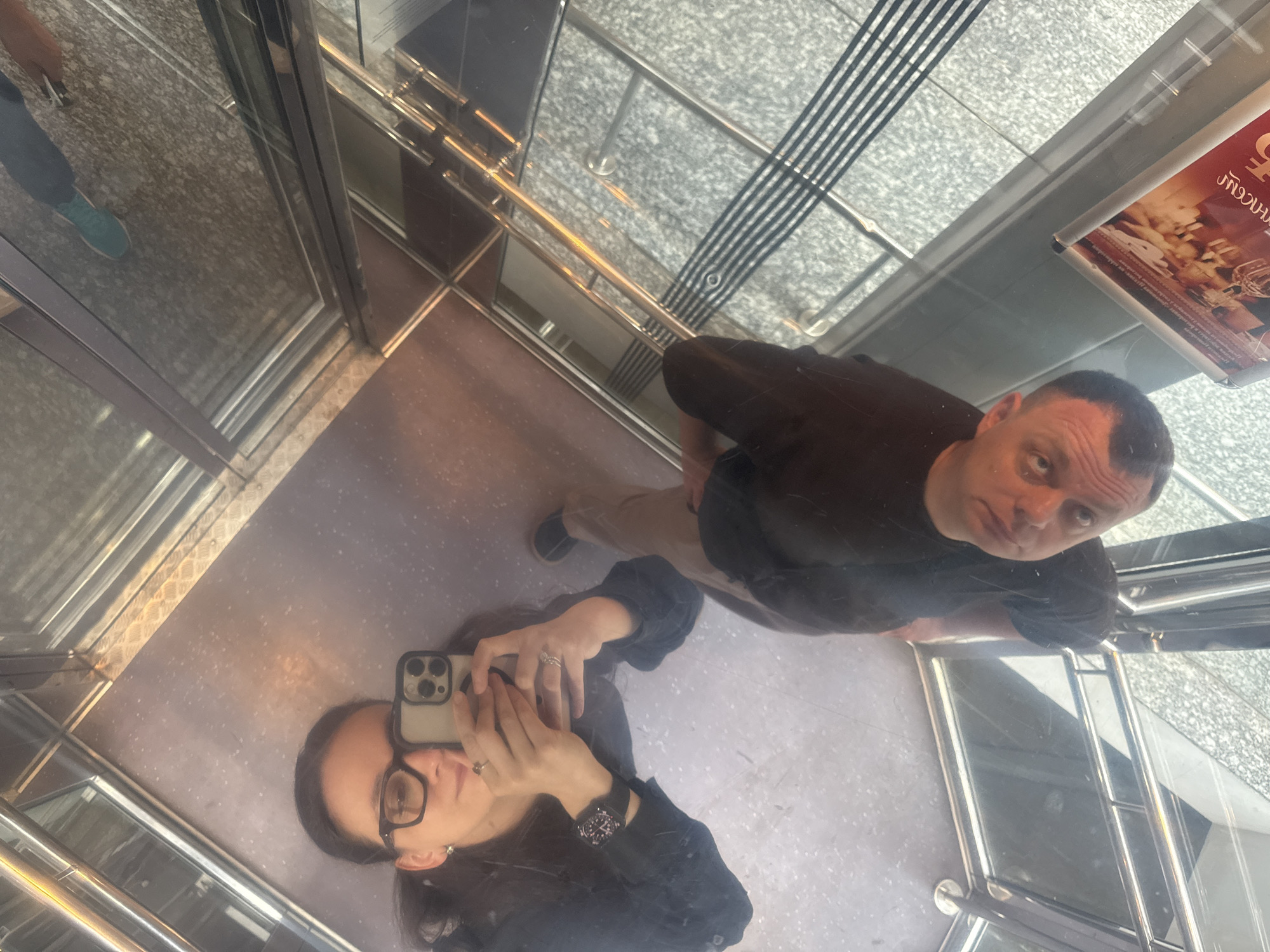 The image shows two people taking a selfie in an elevator. A woman with dark hair and glasses is holding a smartphone and taking the picture. A man with short hair is standing next to her, looking at the camera. The elevator has a mirrored ceiling and walls, creating multiple reflections. The floor of the elevator is a solid color. An advertisement is visible on the wall of the elevator with lettering in Russian. The elevator appears to be in a modern building in Nizhny Novgorod, Russia. The surroundings outside the elevator are visible through the glass walls and ceiling.