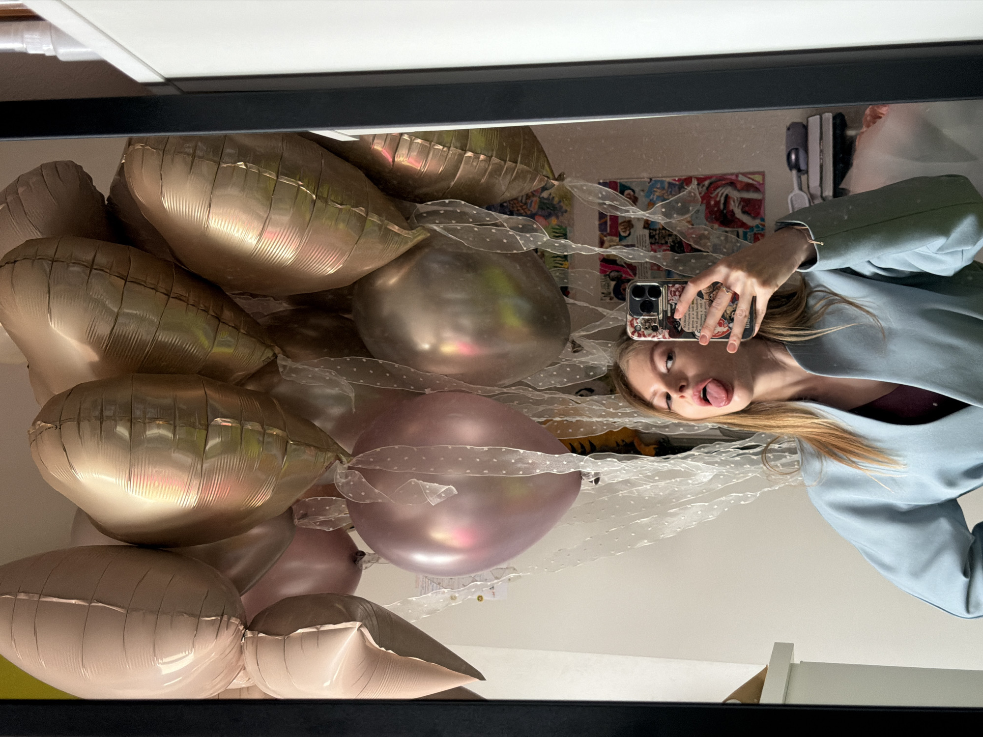 The image displays a playful selfie taken by a young woman reflected in a mirror or a highly reflective surface, oriented vertically. The woman, with blonde hair, is wearing a light blue jacket over a darker top and is captured mid-expression, sticking out her tongue. She holds an iPhone, which has a distinct patterned case featuring small, illegible text and images, in her right hand to take the photo.
The scene is dominated by a large cluster of balloons, suggesting a celebratory occasion. These include numerous metallic, heart-shaped foil balloons in various shades of gold, rose gold, and light pink, intermingled with round, matte light pink latex balloons. Many of the balloons are tied together with white sheer ribbon adorned with small polka dots.
In the background reflection, elements of an indoor setting are visible, including what appears to be wall art or posters and electrical outlets or switches. The lighting suggests an indoor environment. The overall impression is one of festivity and lightheartedness. The location context for this indoor scene is Moscow, Russia.