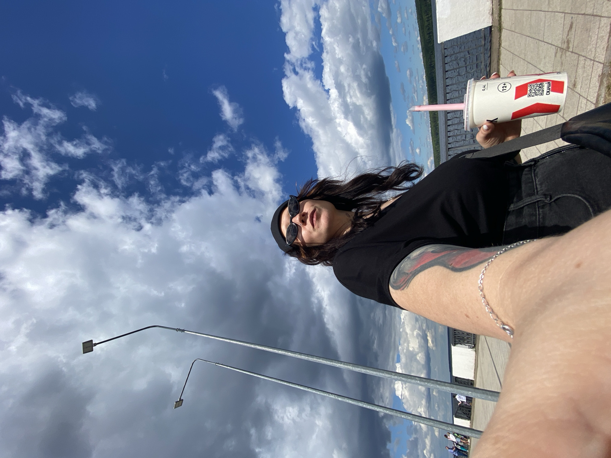 A selfie shows a woman standing outdoors on a paved walkway. She is wearing a black t-shirt, black sunglasses, and a black beanie. She has long, dark hair and a tattoo on her left arm. In her right hand, she holds a white cup with a pink straw. The cup has red markings and text in Cyrillic, including "12+" and a QR code. A black strap from a bag is visible over her shoulder. In the background, two tall lampposts are visible against a partly cloudy blue sky. Beyond the walkway, there is a body of water and a line of trees on the far bank. Several people are walking in the distance along the walkway. The scene appears to be a waterfront promenade in the city of Chaikovsky, Russia, likely during the daytime given the bright sunlight.