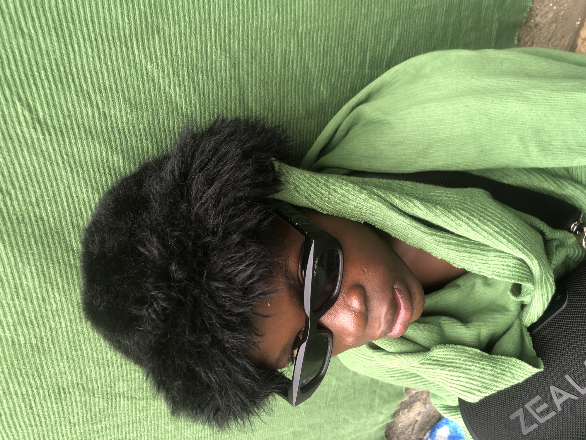 A close-up, high-angle shot shows a young person wearing a black fluffy hat and black sunglasses. The person is draped in a bright green hooded garment, the hood pulled up around their head and neck. The background is a textured green wall, likely outdoors. Part of a black bag with the word "ZEAL" visible is in the lower left corner. The lighting suggests it is daytime. The scene appears to be located in Ado, Nigeria.