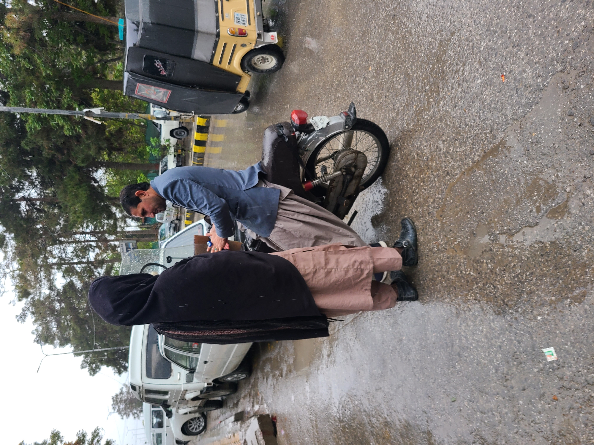 The image depicts a street scene in what appears to be Quetta City Tehsil, Pakistan, given the presence of an auto rickshaw. A man in a blue shirt and shalwar kameez stands next to a motorcycle, facing another person draped in a black cloth or shawl. They seem to be interacting next to a parked white van. The street is wet, suggesting it has been raining. Other vehicles are visible parked along the side of the road. Trees are in the background, indicating a possible urban or suburban location.
