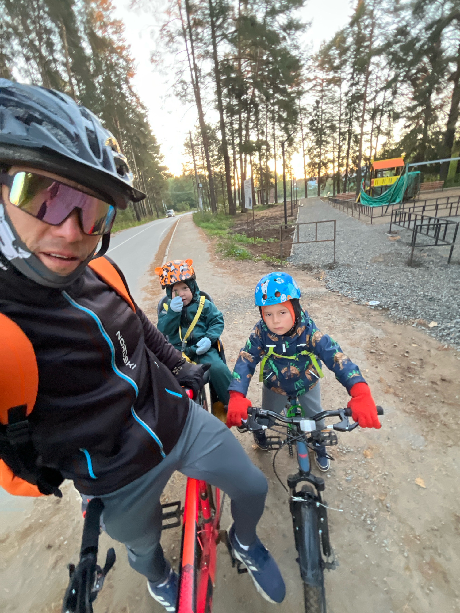 A group of three people are on bicycles in an outdoor setting, possibly a park or recreational area, with trees lining a road. The person in the foreground, wearing a helmet and cycling glasses, is looking at the camera and is on a red bicycle. Behind them, a child wearing a green suit and a tiger-themed helmet is sitting in a child seat on the same bicycle. Further behind, another child, wearing a blue helmet and a dinosaur-patterned jacket with red gloves, is on a separate bicycle. The road appears to be paved, with a gravel path to the right. In the background, there is a playground structure with a yellow and orange slide. The sun is setting, casting a warm glow and long shadows through the trees. The location is identified as Chaikovsky, Russia, based on metadata.