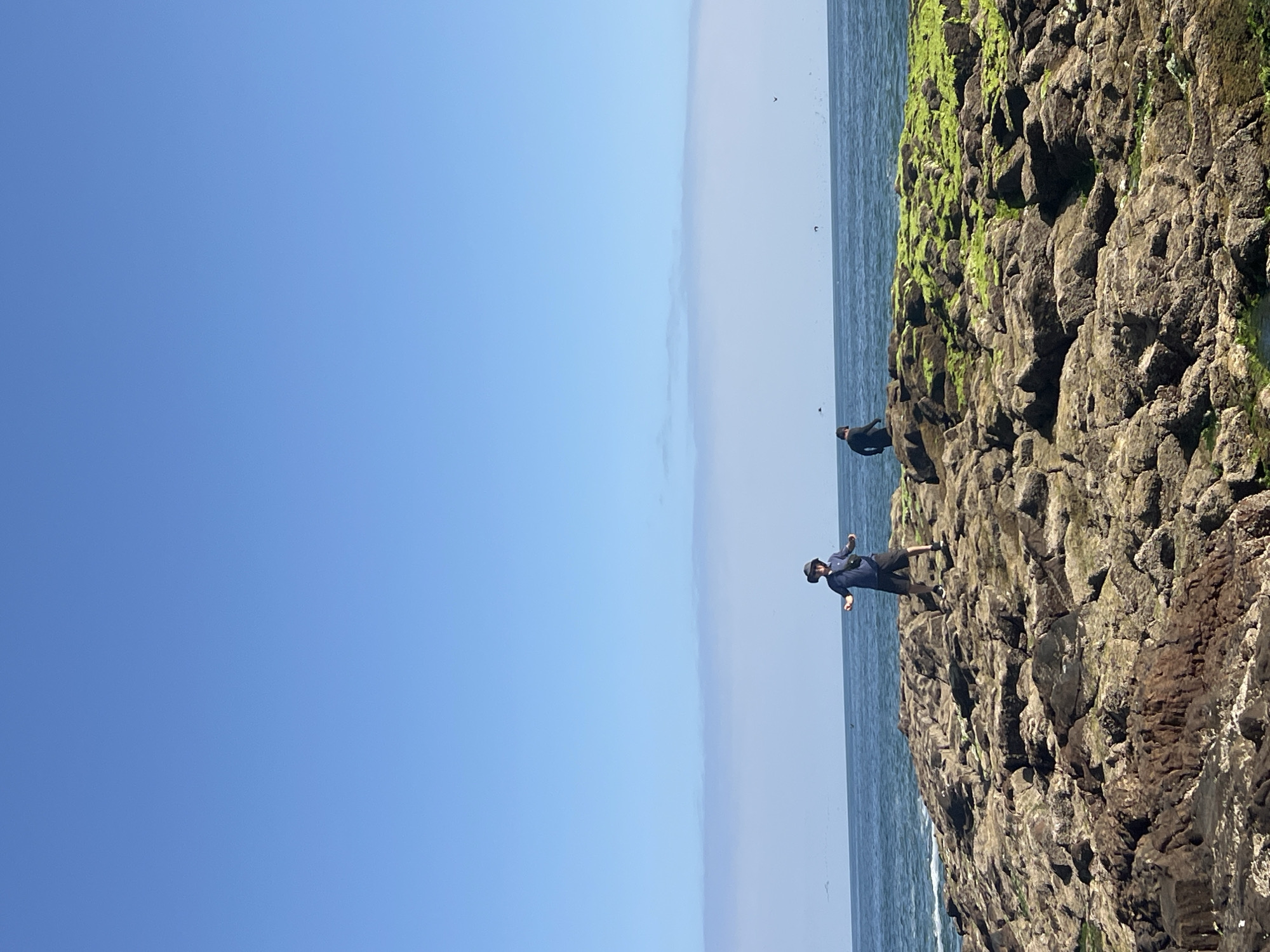 The image depicts a rocky coastal area with two individuals. A man wearing a hat, dark shirt, and shorts is standing on the rocks, extending both arms. Another person in dark clothing is positioned further along the rocks. The rocks are dark and covered in some green moss. The background shows the ocean and a clear blue sky. The location is in the city of Arica, Chile, along the coast.