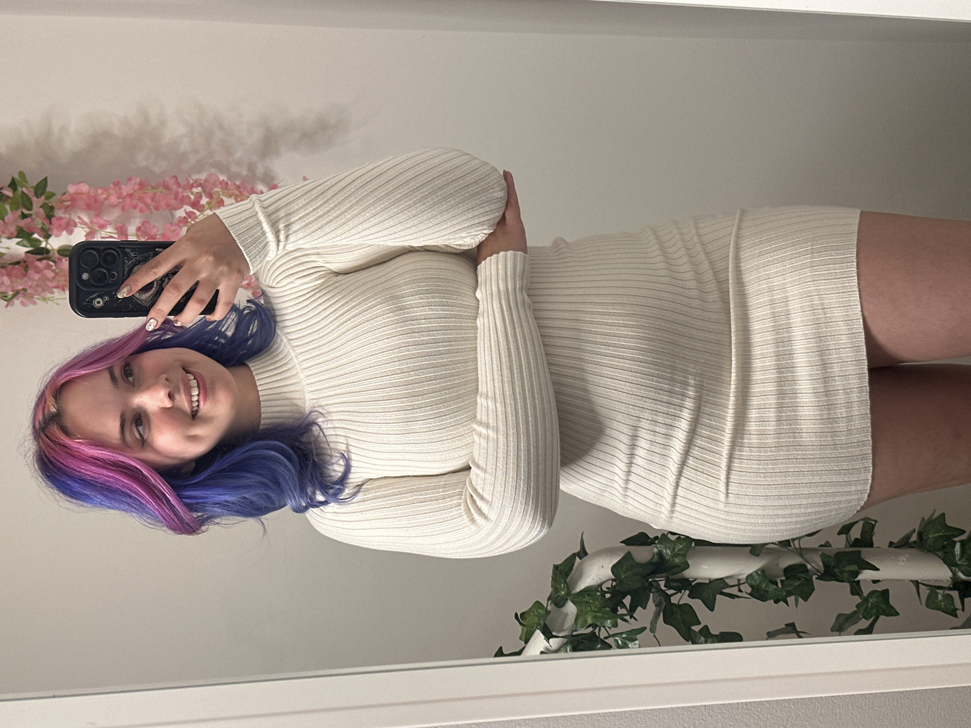 A young woman is captured in a mirror selfie, smiling brightly at the camera. Her distinctive hair features a vibrant blend of pink on one side, transitioning to deep blue or purple on the other. She is wearing a form-fitting, long-sleeved, ribbed knit dress in an off-white or cream color, which extends to her mid-thigh. In her right hand, she holds a black smartphone, likely an iPhone, with a decorative case, positioned to take the photo. Her fingernails have subtle nail art.
The indoor setting shows a plain, light-colored wall as the background. To the upper left, artificial pink cherry blossoms are draped, casting faint shadows. In the foreground, at the bottom of the frame, a strand of artificial green ivy leaves is draped over a white rail or shelf. The overall impression is one of cheerfulness and a casual moment, possibly within a home in the City of Zagreb, Croatia.