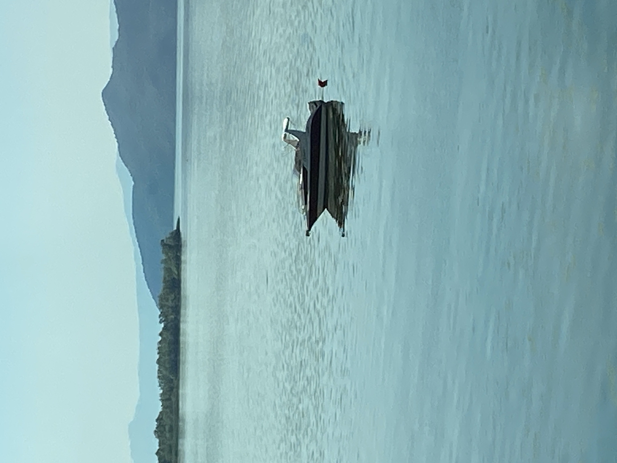 An aerial drone shot captures a tranquil scene in Kruševica, Montenegro. A sleek, dark-hulled motorboat with a white canopy is anchored in calm, turquoise waters. The boat is positioned to the right of the frame, its reflection subtly visible on the water's surface. A small, red buoy floats nearby. In the distance, a lush, green island dotted with trees lines the horizon, transitioning into layers of distant, mist-shrouded mountains. The sky above is a clear, pale blue, creating a serene and expansive atmosphere.