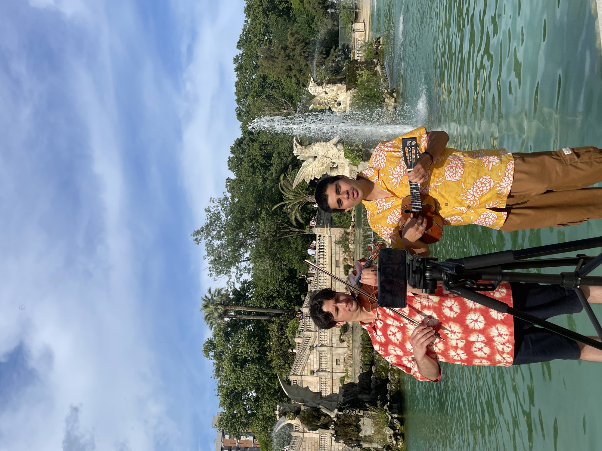 Two men are depicted performing outdoors in front of the Cascada Monumental (Monumental Waterfall) in Parc de la Ciutadella, Barcelona, Spain. The man on the left is of Caucasian descent, with dark hair, wearing a red and white tie-dye short-sleeved shirt and dark shorts. He is holding a violin and a bow, positioned as if playing the instrument, with his gaze directed downwards. A black tripod stands between the two men, holding a smartphone horizontally, likely recording or livestreaming their performance. The man on the right is of South Asian descent, with dark hair, wearing a yellow short-sleeved shirt with white pineapple patterns and brown cargo pants. He is holding a ukulele and his mouth is slightly open, suggesting he is singing or speaking, while looking slightly towards the left.
The background features the elaborate stone architecture of the Cascada Monumental, including a large fountain with turquoise-colored water in the immediate foreground, and ornate stone statues of winged creatures (possibly griffins or dragons) spraying water. One prominent water jet is visible behind the man on the right. Behind the fountain are grand stone staircases, lush green trees, including palm trees, and a distant brick building on the far left. The sky above is blue with scattered white and grey clouds. A single, faint bubble is visible in the upper right quadrant of the sky.