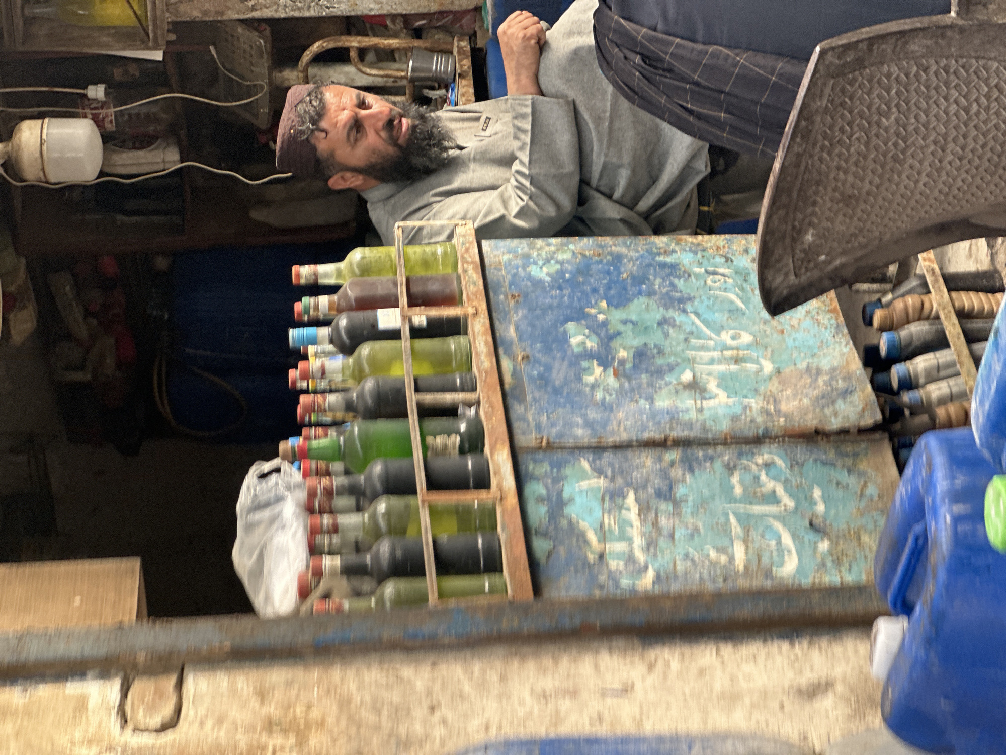 The image shows a man with a beard and a brown kufi cap lying down in what appears to be a cramped shop in Unknown, Unknown. He is wearing a light gray shirt and has his arms folded. To his left, there is a stack of bottles, mostly green and brown, in a metal rack. The rack is placed against a painted blue metal surface with white Arabic script on it. Various objects are visible in the background including an electrical cord, a white lamp, and a blue container. The foreground appears to be a concrete or stone surface.