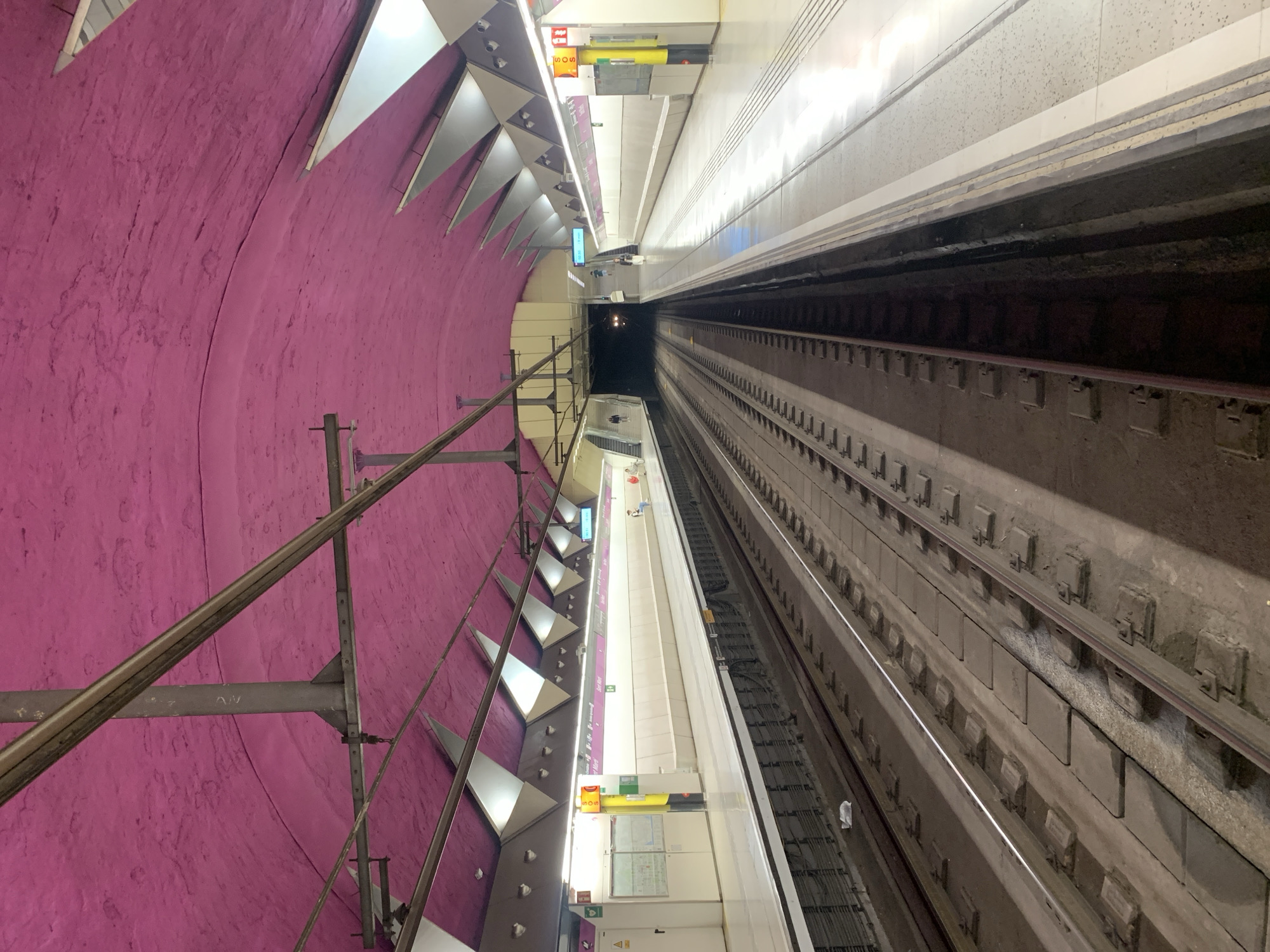 The image displays a modern subway station and tunnel, consistent with a metro system in Barcelona, Spain. The perspective is from the platform looking down the tracks into the distance.
On the left, a large, curved tunnel wall dominates the scene with its vibrant magenta or deep pink color. This wall has a textured finish and features a series of geometric, triangular white panels that project from it, likely serving as light reflectors or decorative elements. A metal railing or support structure runs along the lower part of this pink wall.
The platform, on the right side of the image, is clean and brightly lit by overhead artificial lights, which reflect off its light-colored surface. Visible signage includes a prominent red box with "SOS" written in white, positioned next to a yellow rectangular sign. Further along the platform, smaller green emergency exit signs are present. Text partially visible on a sign along the platform edge reads "Sant Ma...", strongly suggesting "Sant Martí", a known metro station in Barcelona. A few indistinct figures of people can be seen standing on the platform in the distance.
Below the platform and extending into the dark beyond is the subway track system. The tracks are laid on a concrete foundation with various fixtures and components visible between the rails. The tunnel recedes into darkness, with distant lights suggesting its active use and possibly an approaching or departing train. The overall atmosphere is one of a well-maintained, functional, and distinctively designed underground transport hub.