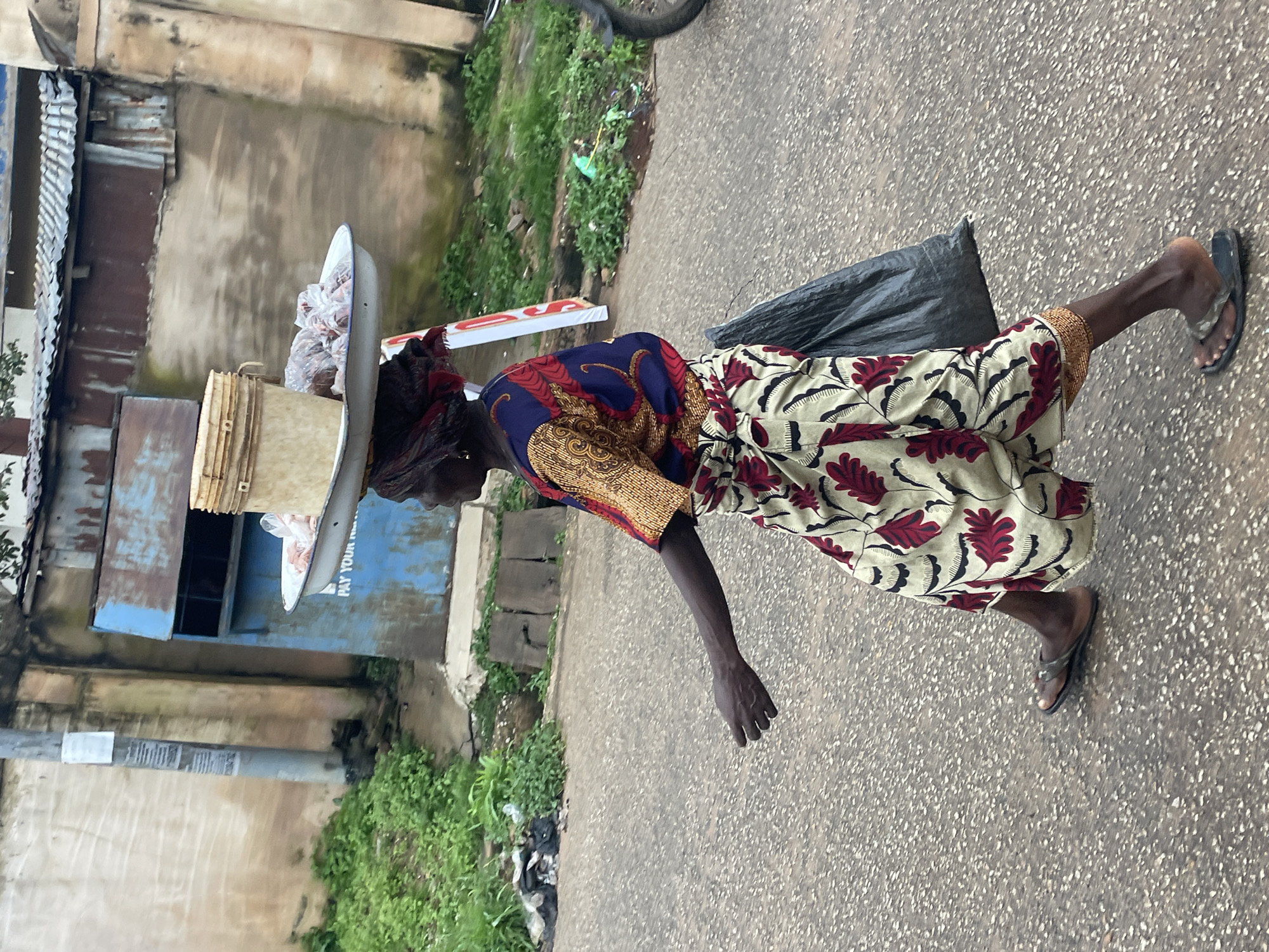 A woman walks along a paved road in Jos, Nigeria, carrying a tray of goods balanced on her head. She is wearing a colorful patterned dress and a headscarf. A large black plastic bag is slung over her left shoulder. In the background, there is a weathered building with a corrugated metal roof and a blue structure that appears to be a kiosk. Green vegetation and scattered debris are visible along the side of the road. A motorcycle is parked to the right of the frame.