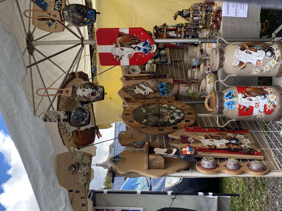 The image displays an outdoor market stall in Abondance, France, identified by the distinctive stone spire of the Abondance Abbey Church visible in the background under a partially cloudy sky. The stall operates under a white canopy/umbrella and is densely packed with handcrafted alpine-themed merchandise.
The foreground is dominated by display grids holding various wooden, ceramic, and metal objects. Prominent items include:
* Numerous wooden plaques and wall hangings of various shapes (rectangular, oval, irregular), many featuring painted portraits of brown and white cows, bulls, chamois, and marmots, often surrounded by white edelweiss and blue gentian flowers. Some wooden pieces are functional, such as oval clocks and a small shelving unit.
* Several painted metal cowbells, hanging from leather straps or displayed on racks, also adorned with cow imagery and flowers.
* Two large, painted metal milk cans standing on the ground, each featuring detailed cow portraits and alpine flowers. One milk can also incorporates a red and white shield design, possibly representing a Swiss or Savoyard canton, with a white cross.
* A red, shield-shaped wall hanging with a prominent white cross and a cow portrait, similar to the Swiss flag motif, is attached to a pole.
* An assortment of ceramic items, including pitchers, teapots, and mugs, decorated with rustic alpine scenes and floral patterns, displayed on shelves or tables.
* Small wooden figures and other decorative objects are interspersed among the larger items.
A dark blue vehicle is partially visible on the left side in the middle ground, parked behind the stall. The ground beneath the stall appears to be covered in green grass or dirt. No people are directly visible in the immediate foreground, but the setup indicates an active commercial environment.