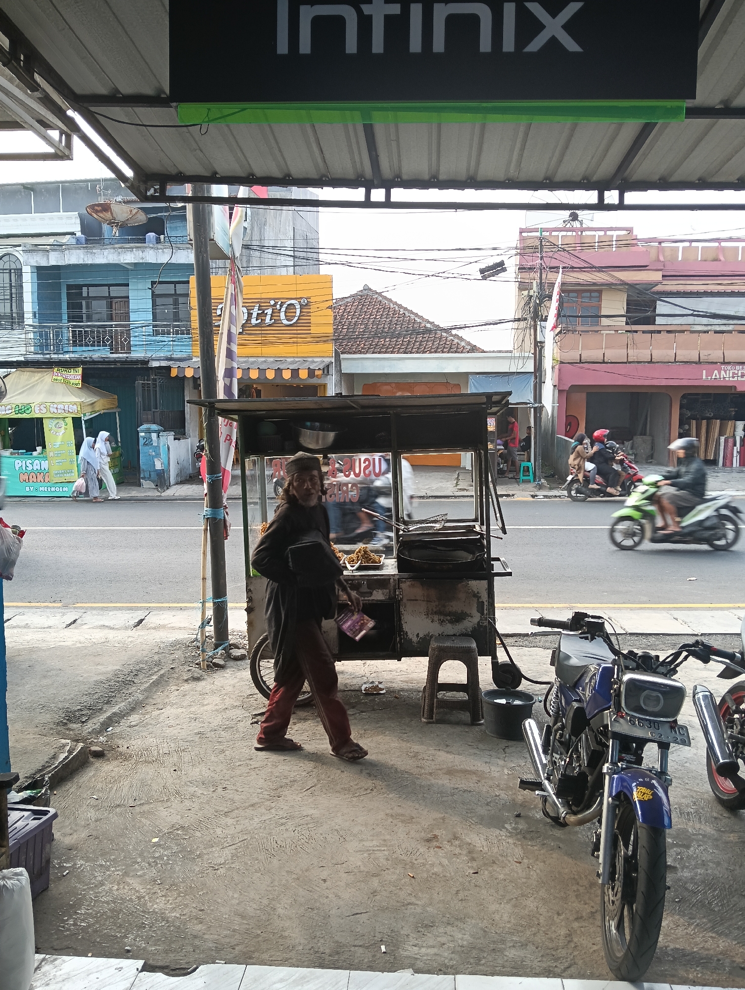 The image depicts a street scene in Desa Cipakat, Indonesia, under an overhang with an "Infinix" banner. A man, appearing to be an elderly street vendor, walks from right to left in the foreground. He is wearing a dark long-sleeved shirt, maroon pants, and sandals, and carrying a dark bag on his left shoulder. He is walking past a food cart which is positioned centrally in the image. The cart has glass panels displaying fried food items and cooking equipment. To the left of the vendor and the food cart, a street with passing motorbikes is visible. On the left side of the street, there is a tent selling "PISAN MAKO ES KRIM" (banana ice cream) with two women in hijabs walking away from it. Buildings line the street, including a blue-fronted building and one with a yellow facade displaying "Opti'O". In the mid-ground, several motorbikes are seen on the road, with one green motorbike in motion on the right. The ground is paved with concrete and some dirt. A blue motorcycle is parked in the lower right corner of the image.