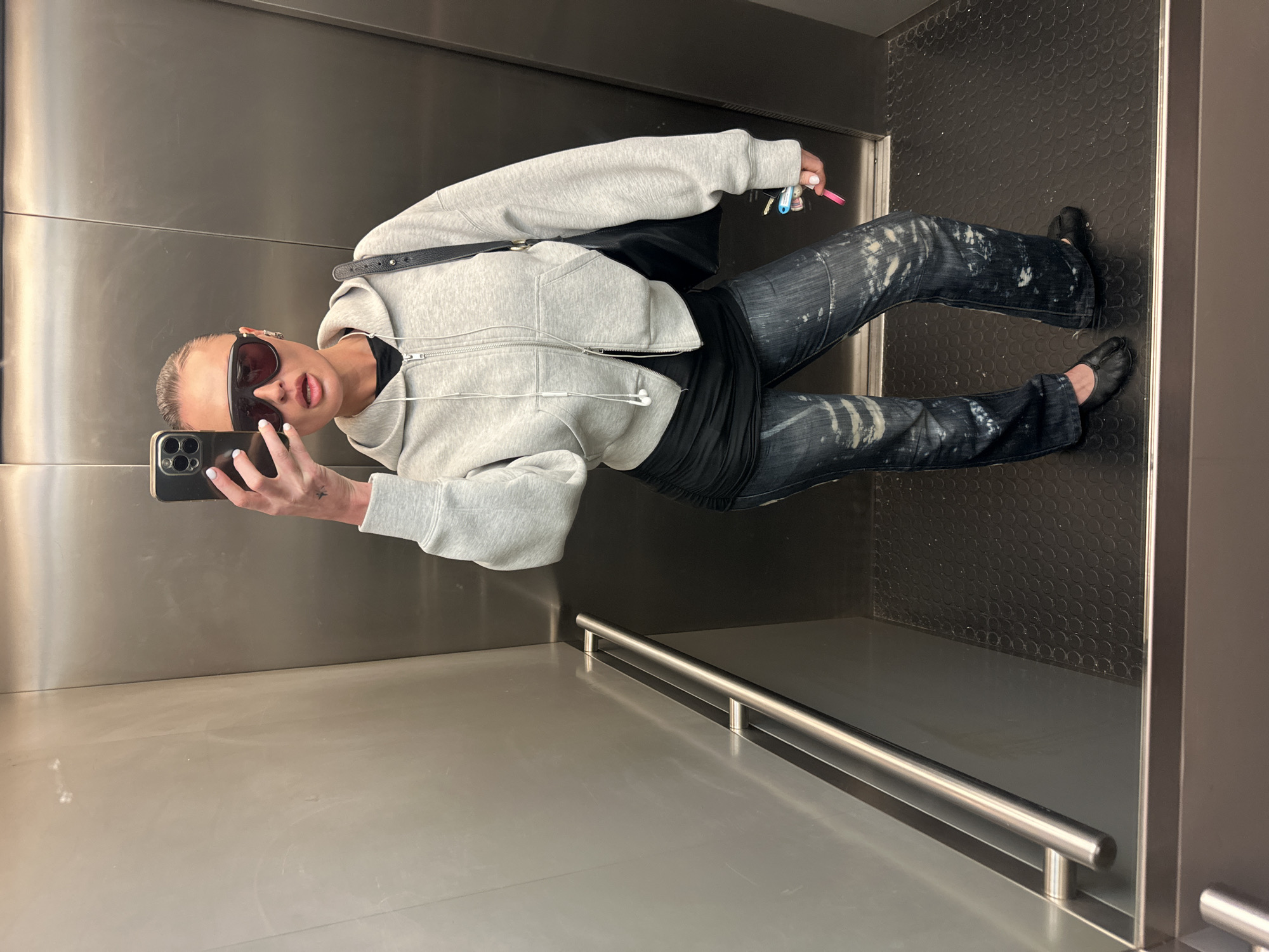 A lone female individual is captured in a full-length mirror selfie within an elevator located in London, United Kingdom. She stands with her body slightly angled, holding a dark-colored smartphone in her right hand to capture her reflection. Her hair is pulled back, and she wears oversized dark sunglasses with a reddish tint. She is dressed in a light grey cropped zip-up hooded sweatshirt over a black long-sleeved top that appears ruched on the left side of the torso. Her dark blue jeans feature prominent white distressed or bleached splatters across the fabric. She wears black flat shoes, specifically Tabi-style with a split toe. A black shoulder bag with a visible strap is worn over her right shoulder. White wired earbuds hang from her neck. In her left hand, she holds a small bundle of keys, including one with a light blue plastic top and another bright pink. A small tattoo, possibly a star or bird, is visible on her inner right wrist. Her fingernails are painted white. The elevator interior features reflective, brushed metallic walls, with a horizontal stainless steel handrail mounted on the left side. The floor is dark, possibly black, with a textured, raised circular pattern. The elevator door, also dark metal, is visible behind her.
