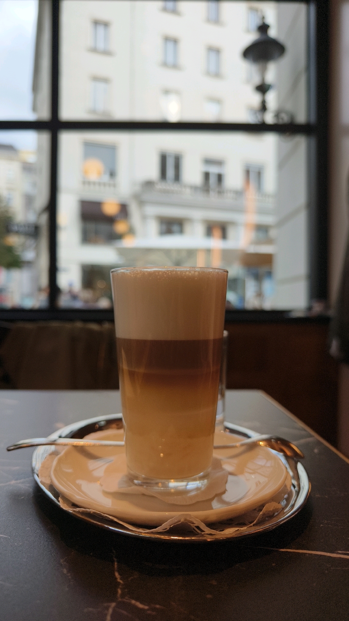 An outdoor scene is captured through a large window of a cafe or restaurant. The window has a dark frame that divides the view. In the foreground, a glass of layered coffee, likely a macchiato or latte, sits on a saucer with a spoon. The coffee is topped with a thick layer of foam. The table surface is dark and textured.
The background visible through the window is blurred, suggesting it is out of focus or a significant distance away. It depicts a European city street with buildings that appear to be in Vienna, Austria. The buildings are light-colored with visible window frames, and architectural details are suggested by the blurred shapes. A street lamp is also visible through the window. The overall lighting is muted, suggesting an overcast day or indoor lighting. There are no discernible people or actions in the frame; the focus is on the coffee and the view beyond.