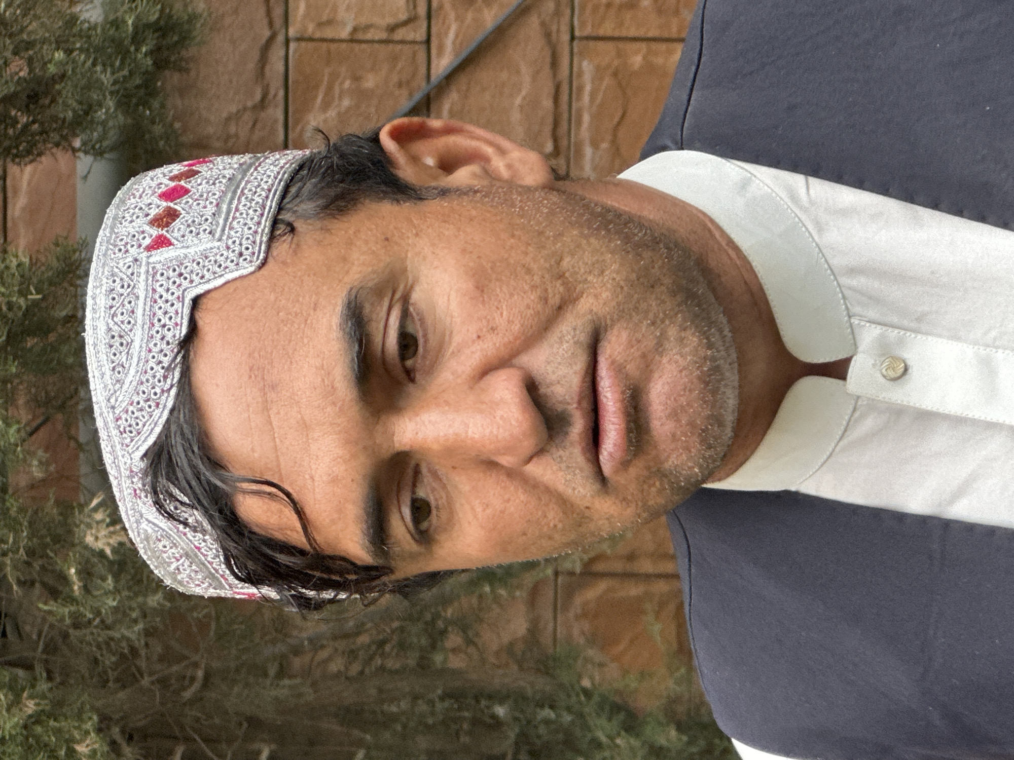 A man is centered in the frame, wearing a white embroidered cap and a collared shirt with a white collar and a dark overshirt. The background features a brick wall behind him, and to the left, foliage appears, suggesting an outdoor setting. The man's facial features are visible, including his eyes, nose, and mouth. The image might be taken in Quetta City Tehsil, Pakistan, although no specific elements within the frame confirm this directly.