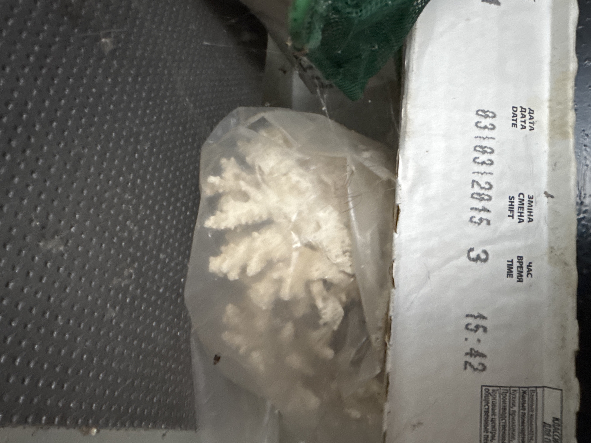 The image shows a close-up view of a white cardboard box with printed text and a plastic bag containing a light-colored, snowflake-shaped object. The box appears to be a product package, possibly for food or a retail item. The text on the box is in Russian and English, indicating "DATE" and "SHIFT/TIME" with corresponding numerical values. The date is "03/03/2015" and the time is "15:42". Below this, there is more text in Russian which appears to be labeling different areas or types of establishments, such as "Жилые поме" (Residential premises) and "Кухни, прихожие" (Kitchens, hallways). The plastic bag contains a white, intricate, branched object that resembles a snowflake or a crystallized structure. The background is dark and textured, possibly a shelf or a surface with a dotted pattern. A green mesh material is visible in the upper right corner of the image. The overall scene is static, focusing on the packaging and its contents. The lighting suggests an indoor setting. There are no people visible. The location is specified as Rozdilna, Ukraine, but there are no specific visual cues in the image that confirm this location.