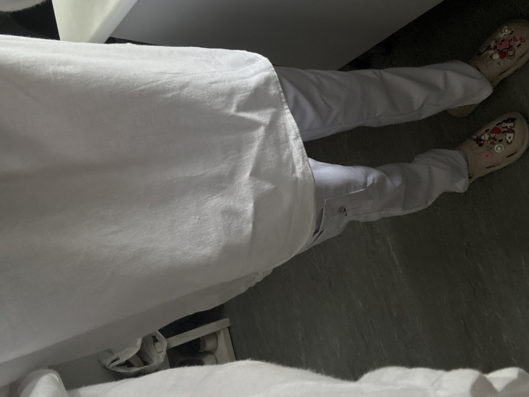 A person wearing white pants and decorated Crocs is lying in bed covered with a white sheet. The location is presumed to be in Saint Petersburg, Russia, but this is not verifiable from the image. The floor is gray.
