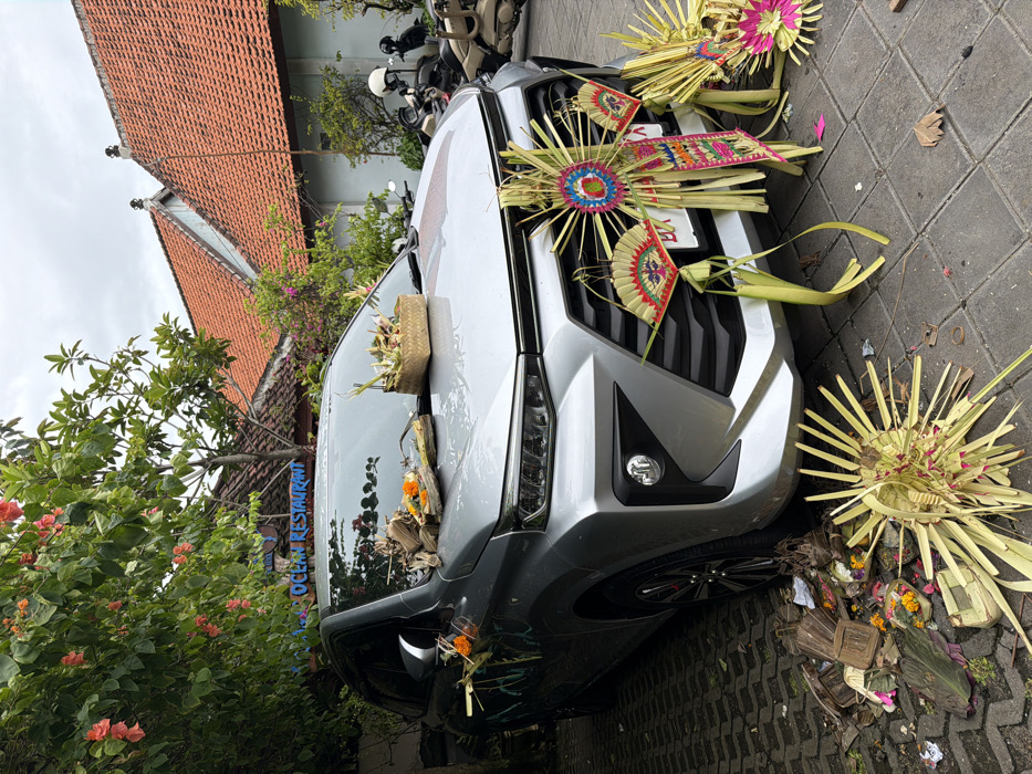 A silver car adorned with Balinese offerings sits on a paved surface. Elaborate palm leaf decorations are attached to the front grill and hood. Baskets of flowers and other materials are visible on the hood and surrounding the front of the car. A building with a red tiled roof is in the background, partially obscured by greenery. Parked motorbikes are also visible in the background.