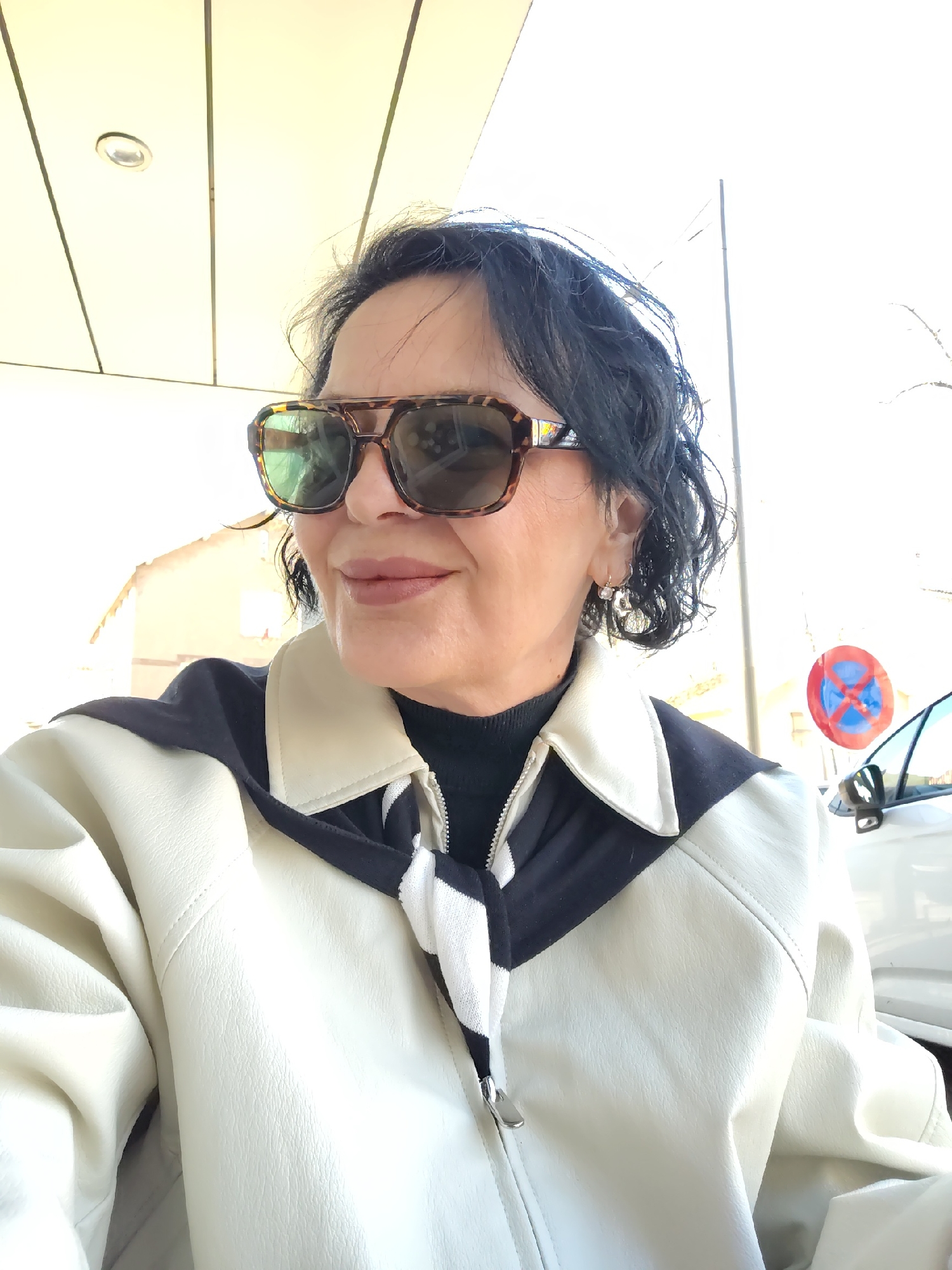 A medium-close-up photograph features a mature woman wearing sunglasses and a cream and black jacket, positioned outdoors. The woman has dark hair and is wearing hoop earrings. Her sunglasses have a patterned frame. The jacket is zipped and has a striped scarf or tie.
In the background, a white car is visible to the right, and a no parking sign is mounted. The scene appears to be in Strasbourg, France, indicated by the design of the no parking sign.