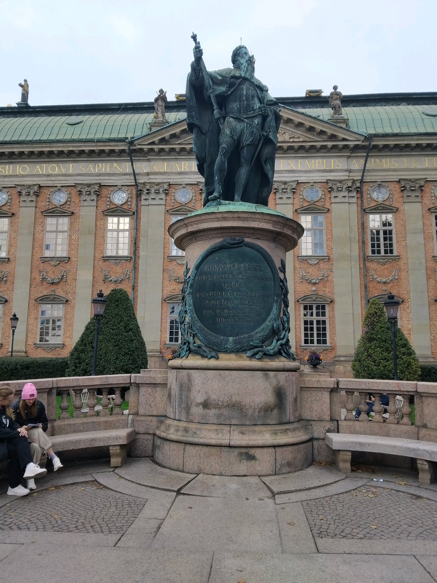 The image shows a statue of a man in historical attire, standing on a stone pedestal in front of a large brick building with a green roof. The building has classical architectural elements, including columns and arched windows. There are also decorative reliefs on the facade. The statue is of King Gustav II Adolf, and the building behind it is the Royal Palace in Stockholm, Sweden. Two people are sitting on a stone bench in the foreground, looking at a piece of paper. The ground is paved with stone tiles and there are some fallen leaves. There are also some bushes and trees visible in the background. The sky is overcast.