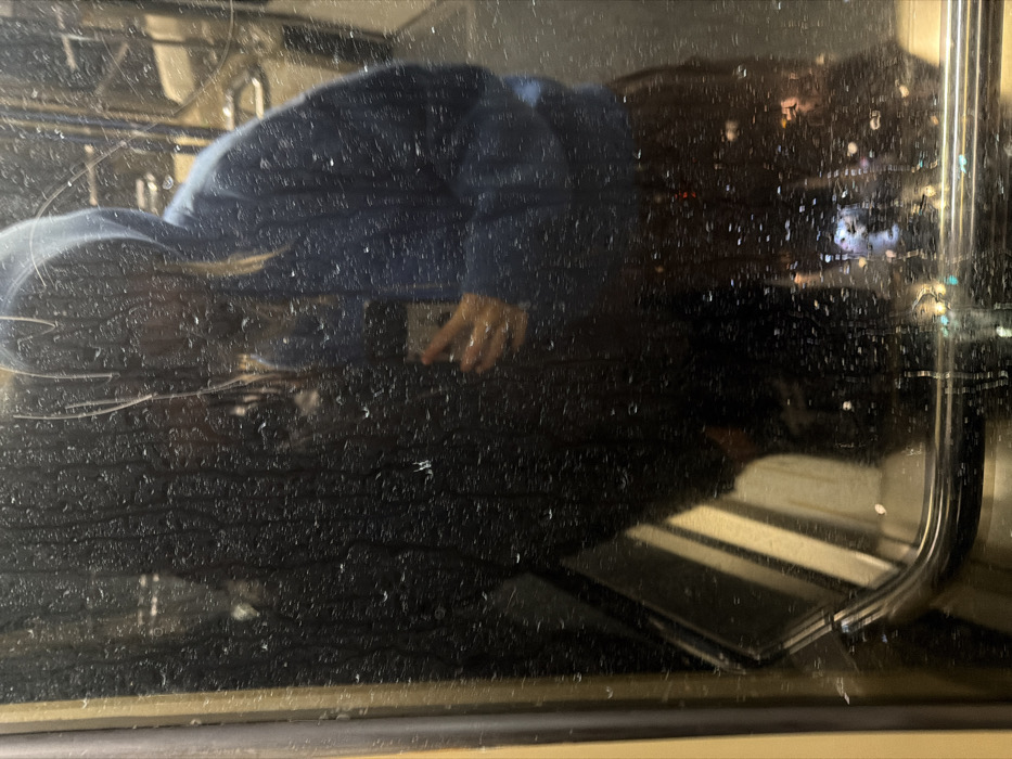 The image shows a blurry reflection in a window, likely on a bus or train. The reflection includes a person wearing a dark jacket and hood. Their face is obscured. A hand is visible, holding a phone. Outside, city lights shine through raindrops on the glass. Inside the bus or train, the metal bar of the seat is visible. The overall impression is one of being inside public transport during rainy weather.