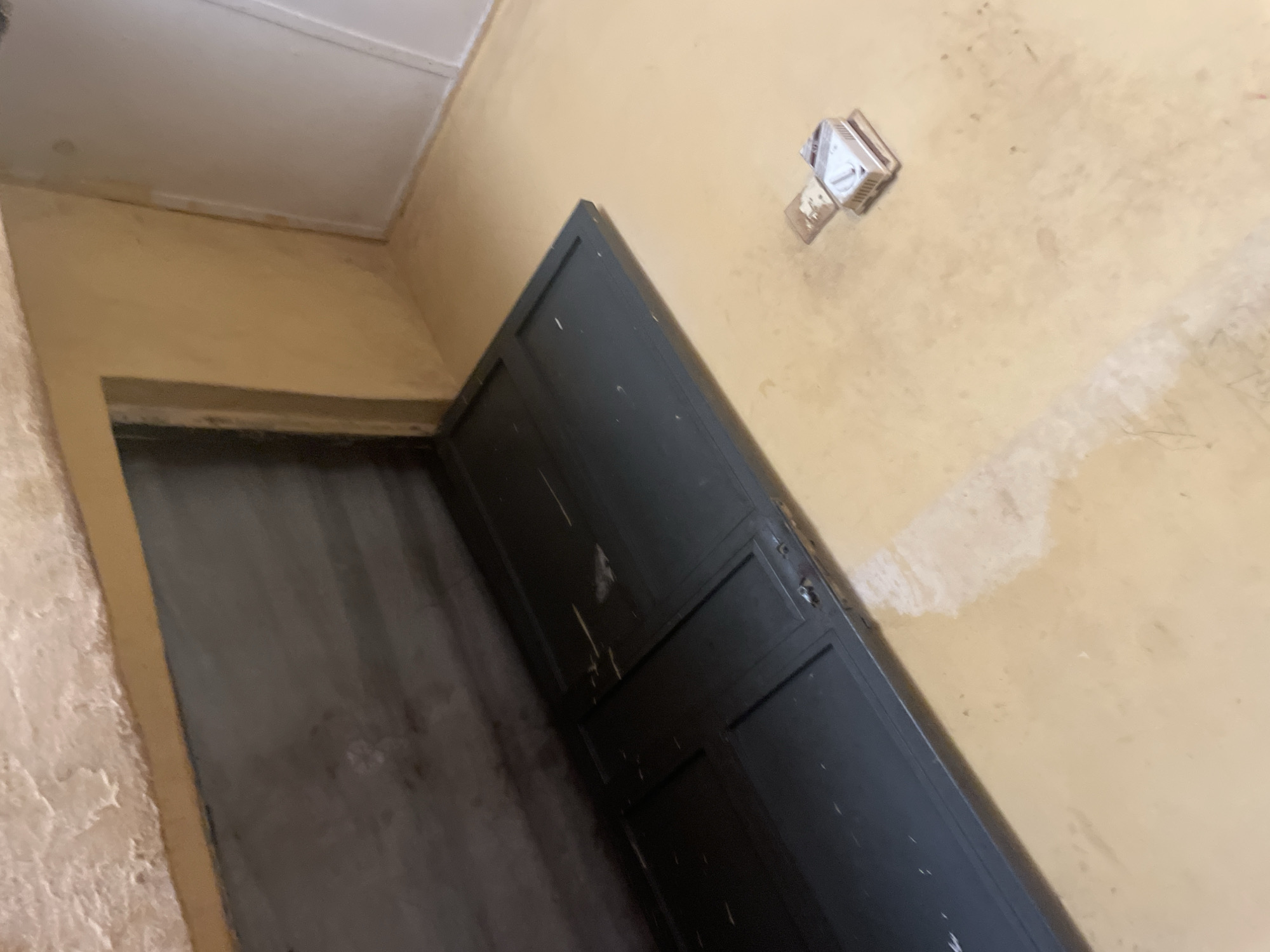 The image depicts a flooding scenario in Lokoja, Nigeria. The scene shows a room with light yellow walls and a light brown floor that appears to be submerged in shallow water. In the foreground, a dark-colored, multi-paneled door lies flat on the flooded floor. To the right of the door, a small, white, and tan electronic device with a dial, possibly a pest repeller or a plug, is visible partially submerged in the water. The background shows a dark, recessed area that might be a fireplace or a niche, with textured material above it. The overall environment suggests a domestic or storage space affected by water damage.