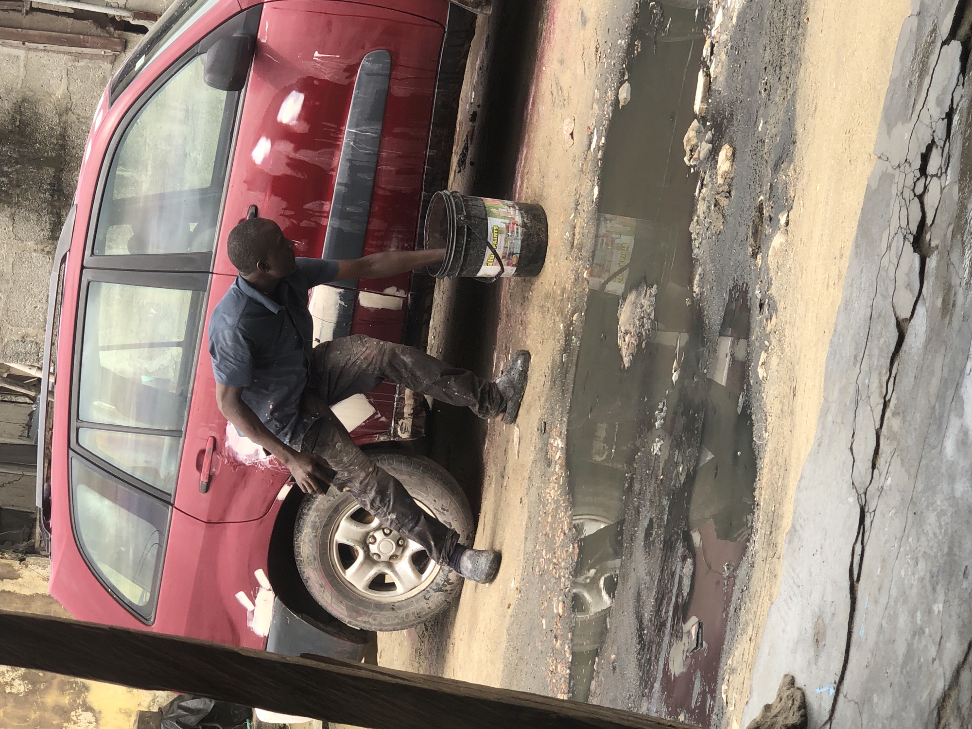 This image depicts an outdoor scene, likely a car repair or bodywork area in Ado, Nigeria. A man, dressed in a dark blue shirt and soiled dark trousers, is seated on the ground next to a red car. He appears to be working on the vehicle, with his right hand submerged in a black bucket, and his left hand near the lower part of the car door. The black bucket has a label that reads "ALL PAINTS" and "PERFECT FINISH."
The red car, possibly an SUV or hatchback, shows visible white patches of body filler or primer on its side panels and door, indicating ongoing repair or preparation for painting. It features black plastic trim along its lower body. The man is positioned near the front wheel, which is a silver alloy type.
The ground is uneven, characterized by a mix of dirt and a large, dark, muddy puddle reflecting its surroundings. Debris is visible within and around the puddle. Further to the right, the ground transitions into cracked concrete. The scene is illuminated by natural daylight, suggesting it's daytime. The man appears focused on his task, engaged in practical labor in a seemingly rugged environment.