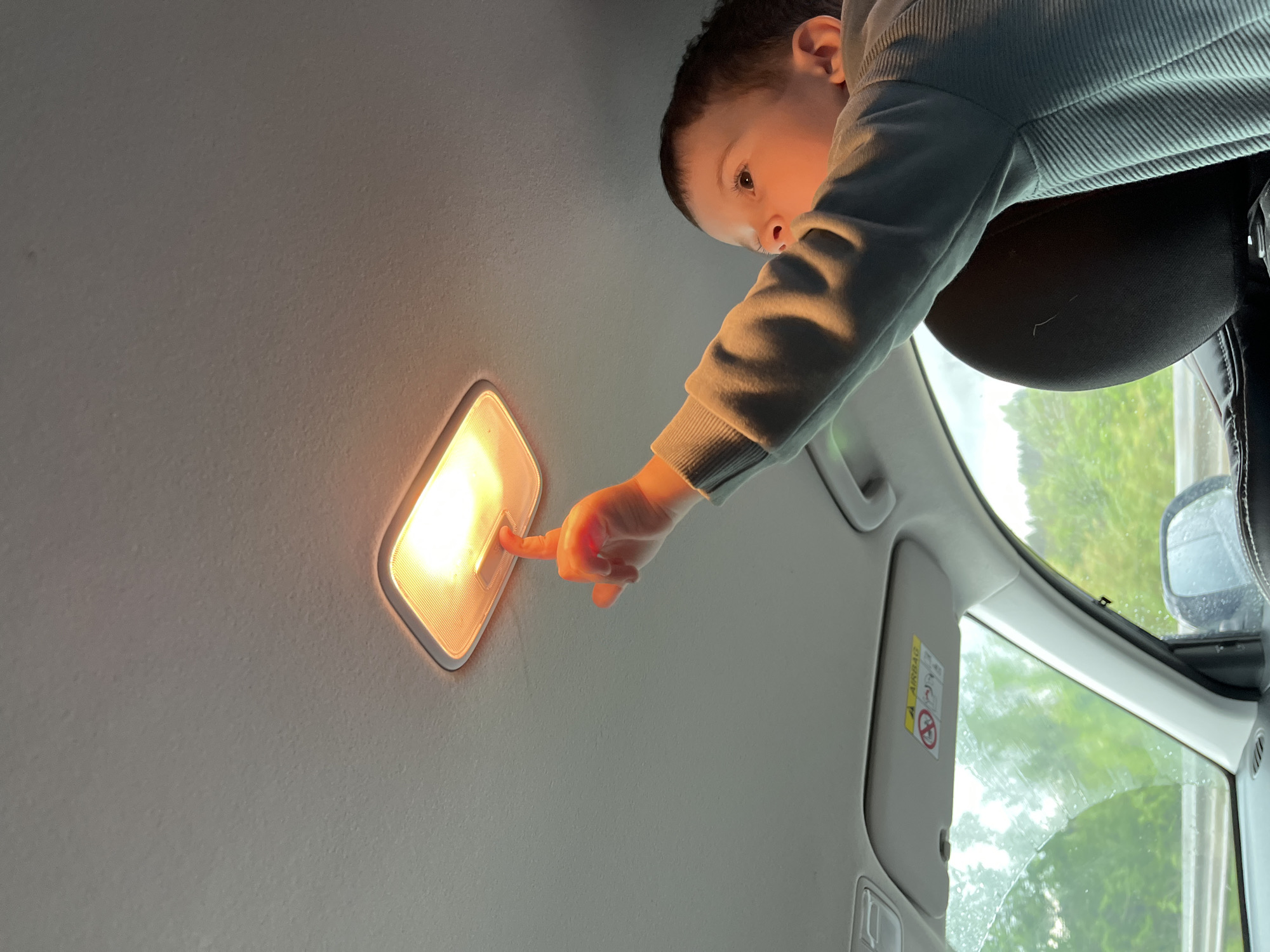The image, taken inside a car, shows a young child engaging with the vehicle's interior dome light. The child, viewed from an upward angle, has dark hair and wears a light grey or blue ribbed long-sleeve shirt. Their left index finger is pressed against the warmly illuminated, rectangular light fixture embedded in the car's grey ceiling, casting a glow on their hand and face. The child's eyes are focused intently on the light, suggesting curiosity or playfulness.
To the right, a portion of a black car seat headrest is visible. Below it, through the car window, a blurred outdoor scene features green foliage, indicative of trees or bushes. The window surface appears to be wet with raindrops or condensation, suggesting overcast or rainy weather conditions. A light-colored sun visor, partially visible on the right side of the frame, displays a yellow warning label with the prominent text "AIRBAG" and a graphic indicating the danger of placing rear-facing child seats in front of active airbags. The scene captures a moment of childhood exploration within the confines of a car interior.
