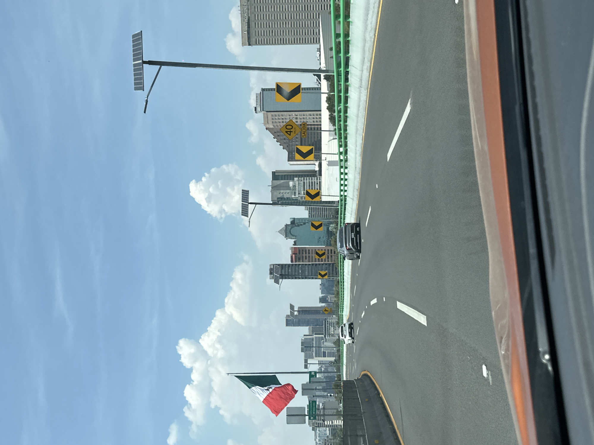 The image shows a highway in Mexico City, Mexico. The road is asphalt with white lane markings and a yellow line along the shoulder. A green metal guardrail is visible on the left side. Several skyscrapers are in the background, interspersed with green trees and vegetation. Signs with downward-pointing arrows and a 40 km/h speed limit sign are placed along the roadside. Solar panels are installed on poles beside the highway. The Mexican flag is prominently displayed in the lower left of the frame. Cars are on the road. The photo was taken from inside a vehicle, with part of the car's frame visible on the right side of the image. The sky is blue with scattered clouds.