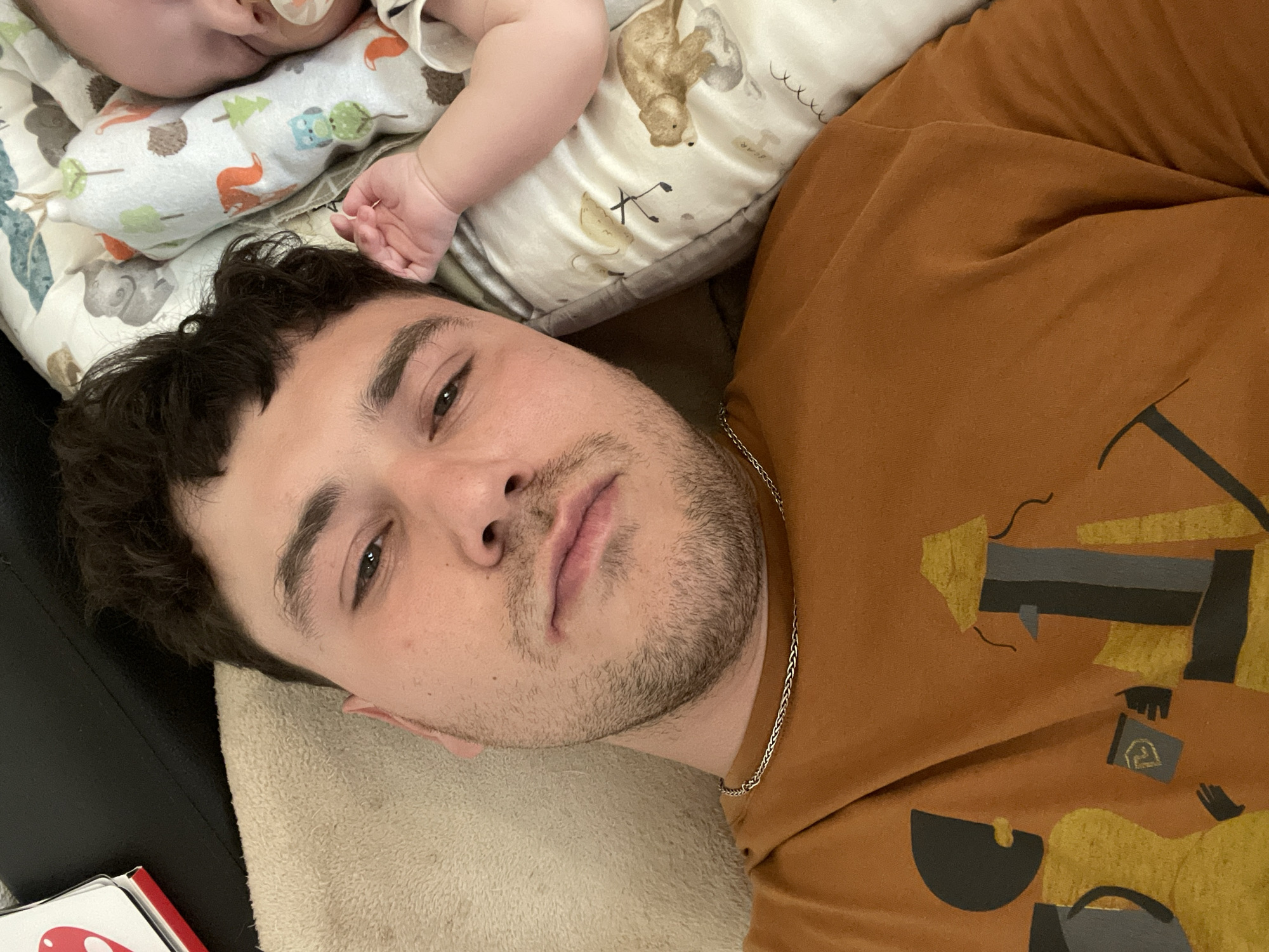 A selfie taken indoors shows a man lying on his back, with a baby partially visible near his head. The man has short, dark curly hair, light skin, and a beard. He is wearing a brown t-shirt with an abstract design and a silver chain around his neck. The baby is lying on a patterned pillow. A black object is visible to the left, along with the corner of a red and white object. The location of the photograph is not indicated. The city of Bar, Montenegro is not relevant to the image.