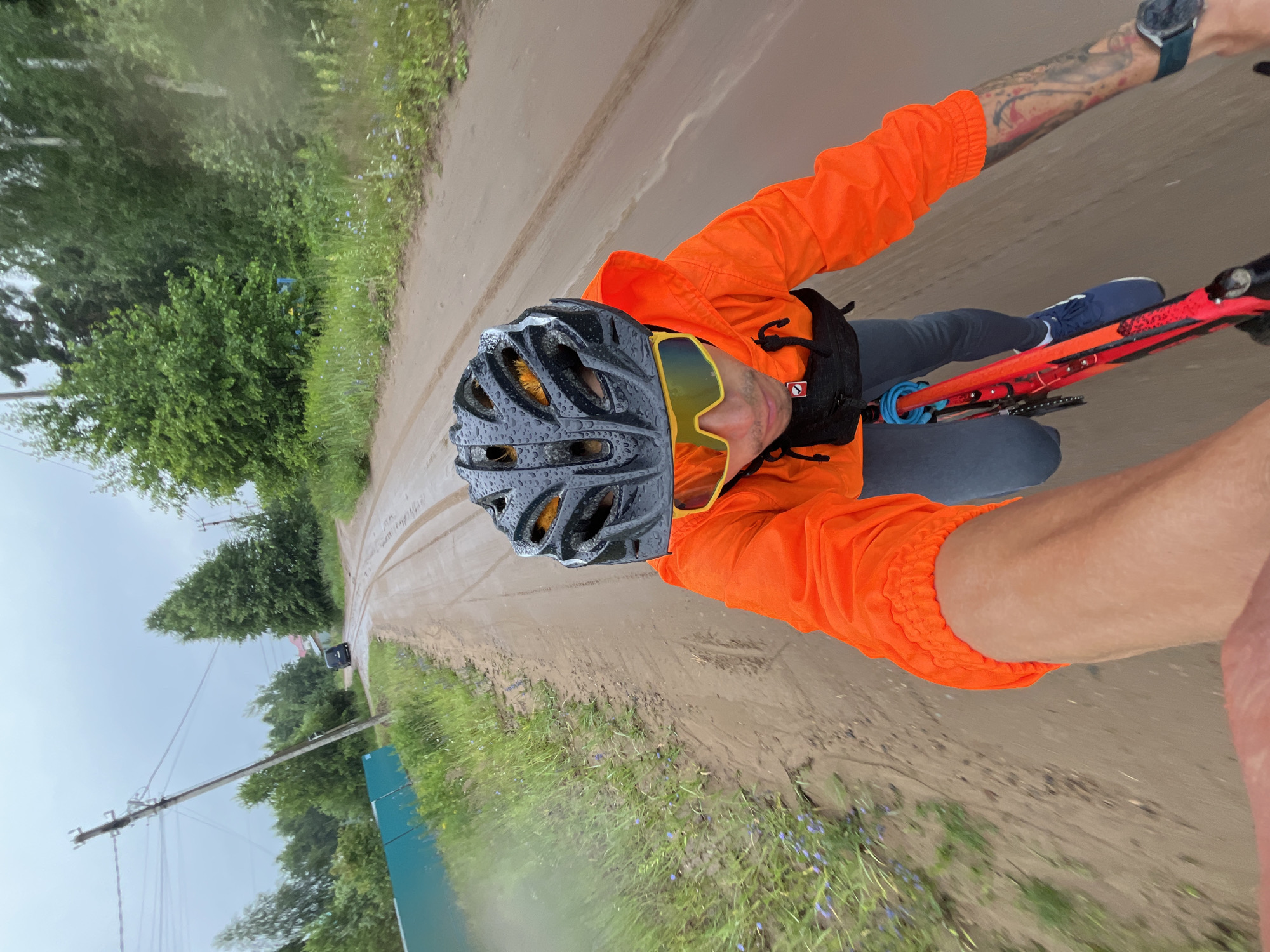 This is a first-person perspective media file showing a person riding a bicycle on a dirt road. The person is wearing an orange jacket, a black helmet with water droplets, and yellow sports sunglasses. They also have a watch on their left wrist and tattoos on their left arm. The bicycle is red and black. The road is muddy and wet, indicating that it has been raining. The surrounding scene includes lush green trees and grass, with some small purple wildflowers visible. There are utility poles with wires and a green fence in the background. The sky is overcast, suggesting a cloudy or rainy day. The overall mood is one of outdoor activity, likely a bike ride in challenging weather conditions. The location is identified as Chaikovsky, Russia. No visible text is present in the image.