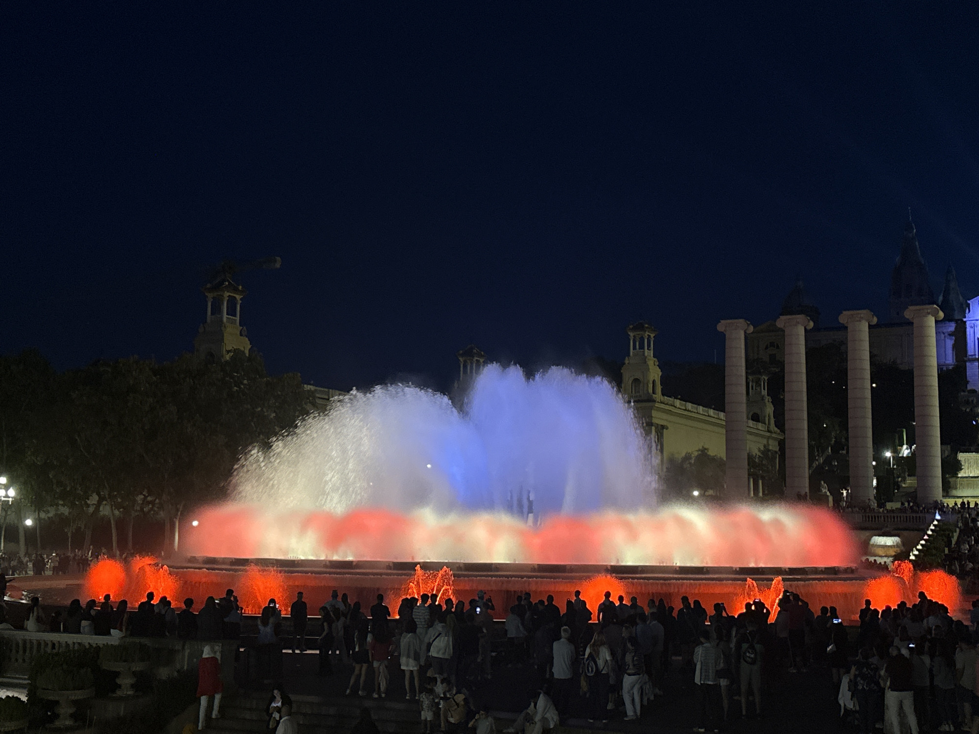 This nighttime image captures the Magic Fountain of Montjuïc in Barcelona, Spain, during a synchronized light and water show.
In the center, a large, multi-tiered fountain displays vibrant illumination. The main water jets project upward in a broad spray, illuminated predominantly in white on the left and blue on the right, creating a distinct two-toned column of water. Below these primary jets, a wider expanse of water features a flowing pattern, intensely lit in various shades of red and orange. Smaller, localized eruptions of water, also red-orange, are visible closer to the foreground.
A dense crowd of people is gathered around the fountain, primarily silhouetted against its bright lights. Spectators are positioned on multiple levels, including a wide platform directly in front of the fountain and on steps and ledges in the immediate foreground. Many individuals are holding up smartphones, indicating they are likely photographing or videoing the display.
In the background, architectural elements characteristic of the Plaça d'Espanya area are visible. To the right and elevated, classical columns and sections of the Palau Nacional (National Palace) are illuminated. To the far left, a distinct, ornate tower structure is partially visible above trees, suggesting one of the Venetian Towers or a similar monument. Other buildings with arched windows and domed roofs are also present in the mid-ground. Trees line the left side of the scene, partially obscuring other structures. The sky above is a deep, dark blue, typical of twilight or night.