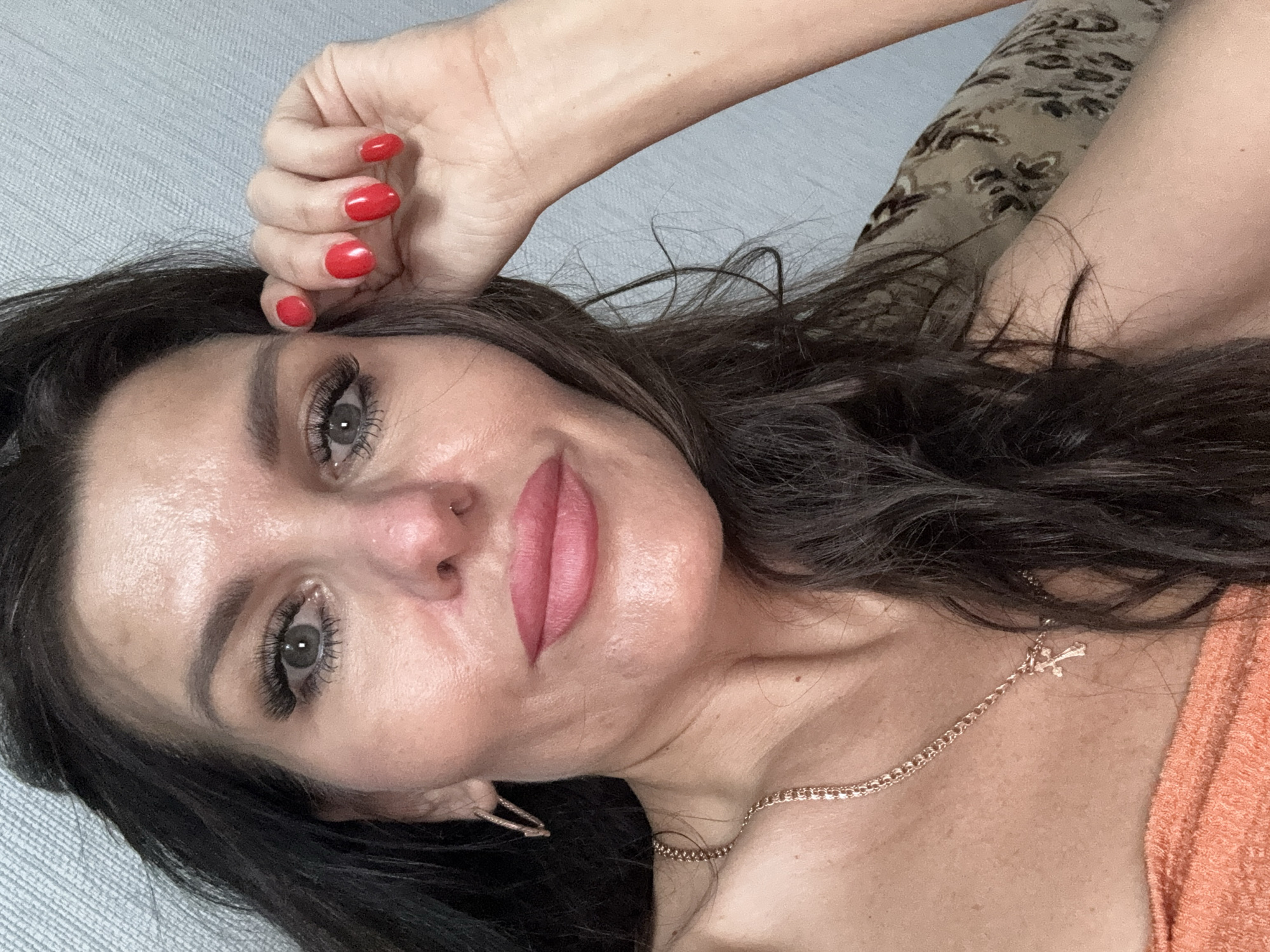 A close-up selfie captures a woman with dark, wavy hair and fair skin, positioned indoors against a light blue, textured wall. She is looking towards the camera with a neutral expression, her head tilted slightly. Her right hand, adorned with bright red nail polish, is raised to her forehead, with her fingers gracefully resting on her temple. The woman is wearing subtle makeup, including mascara, eyeliner, and pink lipstick. A delicate gold necklace with a cross pendant is visible around her neck, and a hoop earring is in her left ear. Her attire includes an orange, textured top and a patterned fabric, possibly a couch or blanket, behind her. The image does not depict any specific location within the city of Dubovaya, Russia.