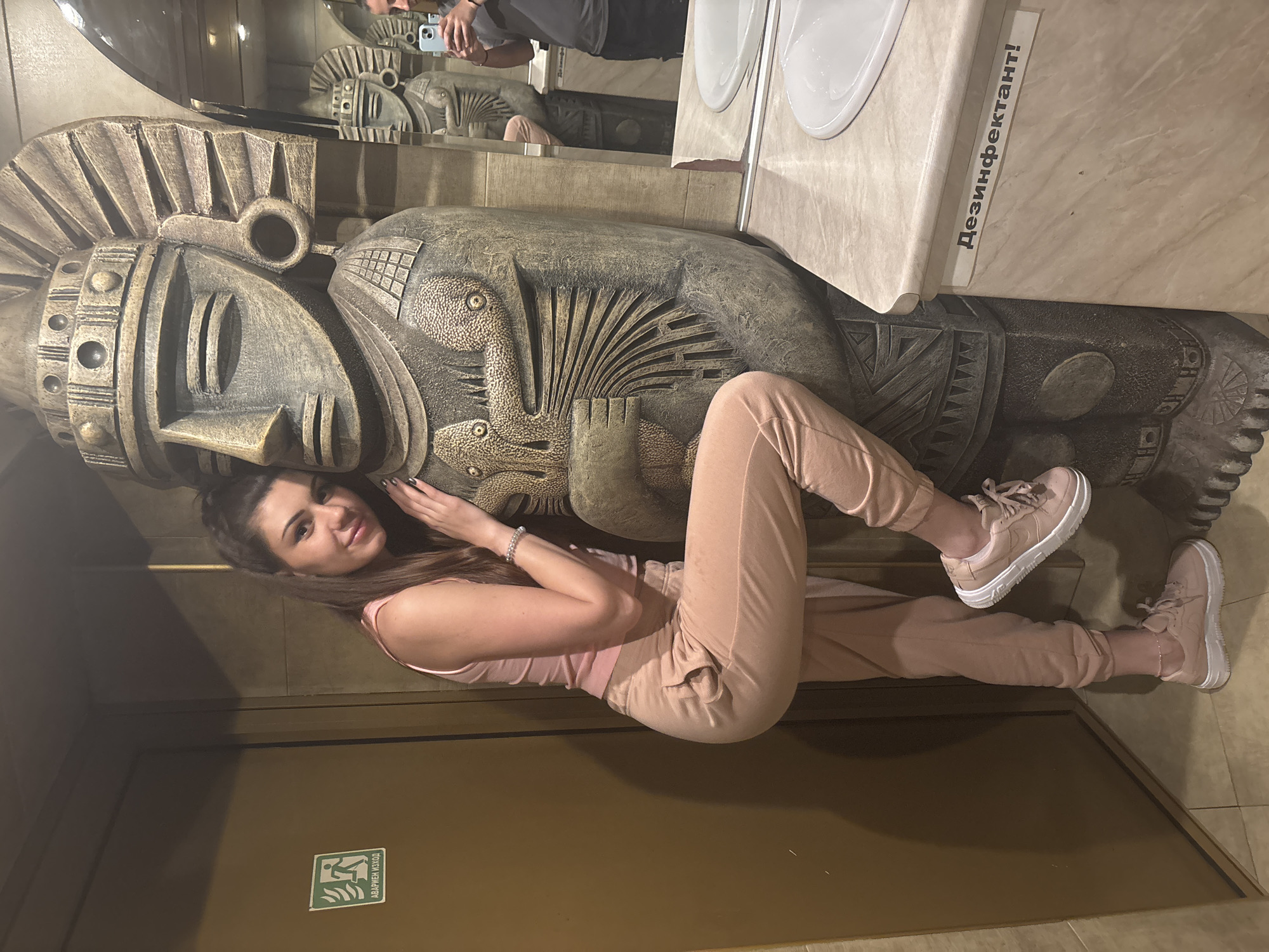 A young woman poses with a large sculpture in a bathroom, possibly at a restaurant or public space. The woman, wearing a pink top and sweatpants with matching sneakers, rests her head near the face of a large, weathered, brown statue. The statue appears to be an anthropomorphic figure with intricate detailing on its headpiece and torso. The bathroom features a sink and a mirror, with a person visible in the reflection taking a photo. A sign in Russian on the right edge of the photo states that the surface is "Disinfected!" In the lower left corner is a brown door with an emergency exit sign in Russian. The location appears to be in Sofia, Bulgaria, based on the Russian Cyrillic signage.