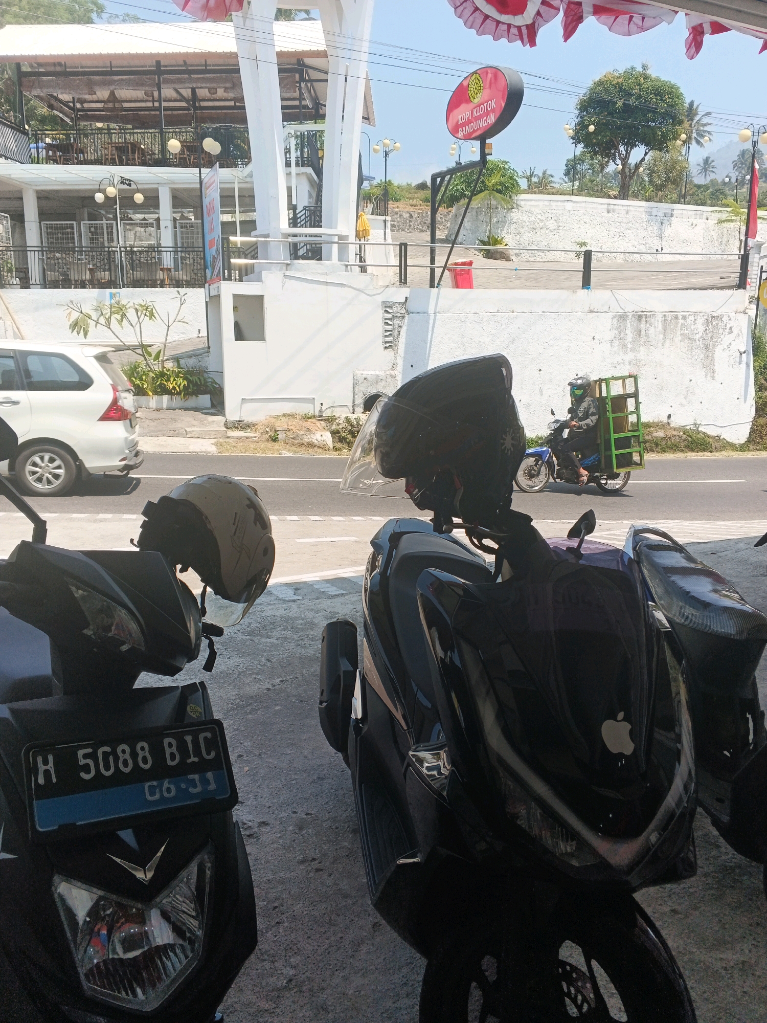 In Pakopen, Indonesia, on a sunny day, a street scene unfolds with two parked motorcycles in the foreground. The motorcycle on the left displays a license plate with the characters "H 5088 BIC" and the characters "06.31". A helmet rests on the handlebars of this motorcycle. To the right, another black motorcycle is visible, with an Apple logo emblazoned on its front. In the background, a person wearing a helmet and a jacket rides a blue motorcycle, carrying a green crate on the back. A white van is parked on the side of the road. Beyond the road, a white building stands with a restaurant situated on an upper level. A circular red sign with yellow lettering reads "KOPI KLOTOK BANDUNGAN". The environment is characterized by white walls, trees, and a clear sky.
