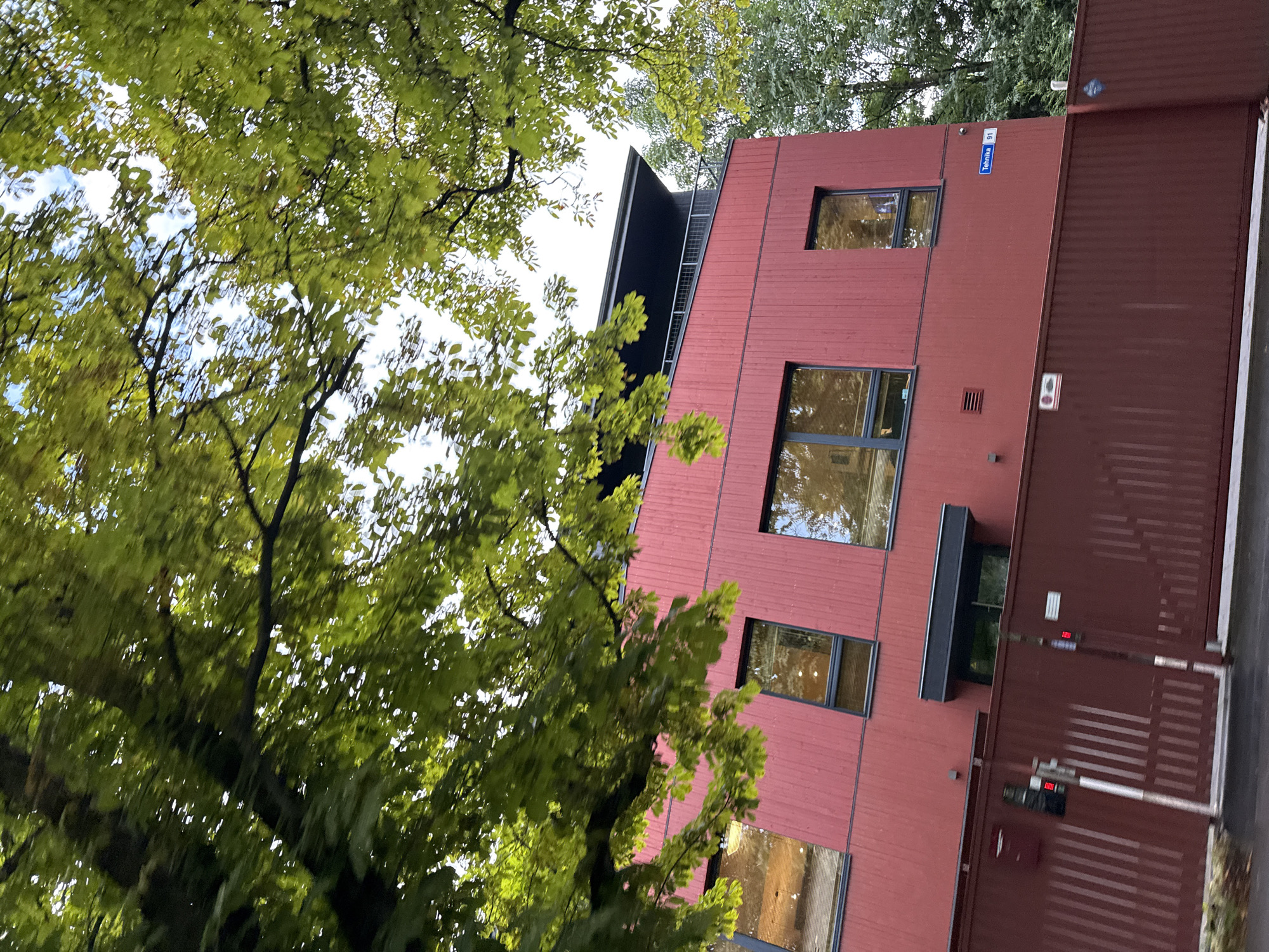 This is a medium-distance, slightly angled exterior shot of a modern red building with horizontal wood siding, partially obscured by green trees in the foreground. The building features several large, dark-framed windows that reflect the surroundings. A dark-colored balcony with a railing is visible on the upper level. In front of the building is a tall, dark red metal fence. A street sign reading "Tehnika 91" is mounted on the building's exterior wall. The scene appears to be in Tallinn, Estonia, based on the street sign. The sky is visible through the leaves and is overcast.