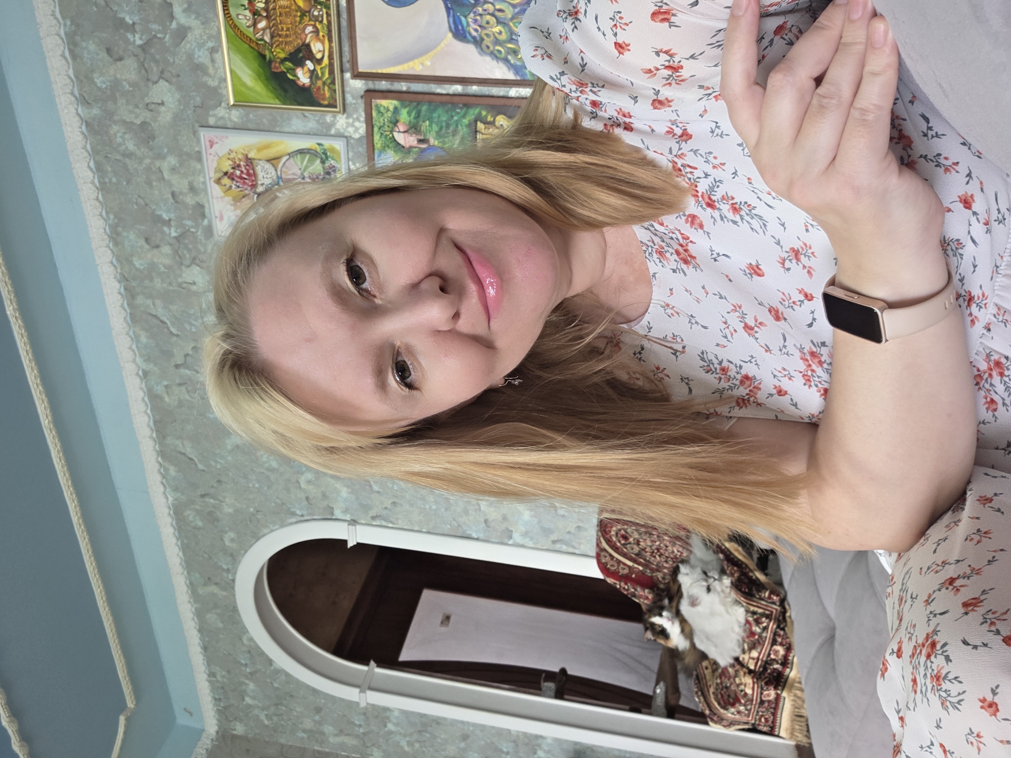 The media file shows a portrait of a blonde woman with light skin and rosy lips, wearing a white floral dress. She has a watch on her left wrist. The image is taken in a room with blue walls and decorative paintings on the wall. There is an arched doorway with a patterned rug and a cat visible in the background. The lighting suggests it is daytime. The woman is looking towards the camera with a slight smile, conveying a relaxed and pleasant emotion. There is no visible text in the image. The overall setting appears to be domestic.