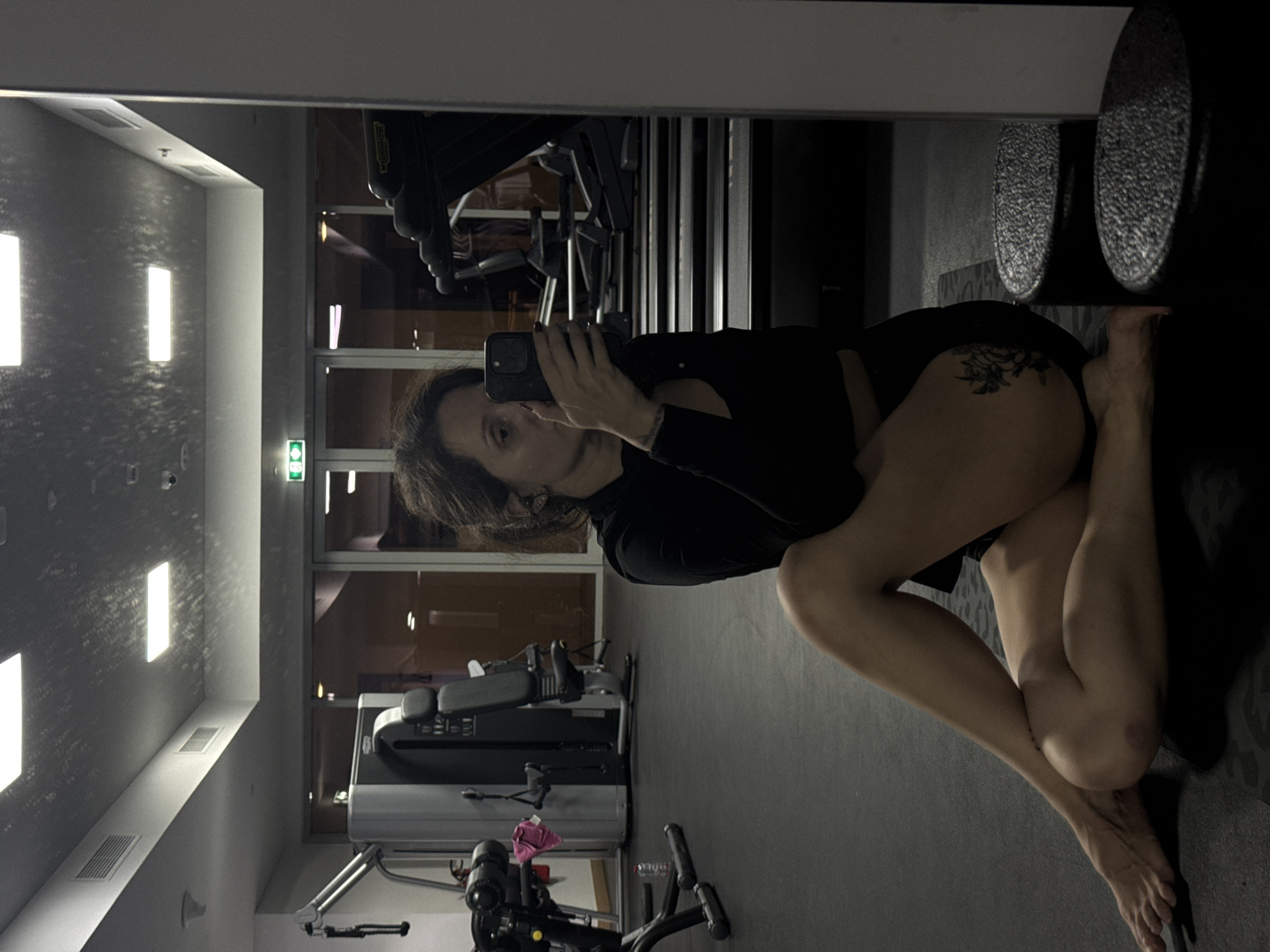 A woman is captured in a mirror selfie within a gym or fitness center in Dubai, United Arab Emirates. She holds a dark-colored smartphone to take the photo, partially obscuring her face. She has dark hair and is wearing a black long-sleeved top and black shorts. A floral or organic-patterned tattoo is visible on her left hip/upper thigh. She is seated or kneeling on an exercise mat with her legs bent and crossed, and her bare feet are visible.
The indoor setting features light-colored, possibly textured walls illuminated by bright rectangular overhead lights. Large windows in the background reveal a dark, nighttime view, indicating the photo was taken after dusk. Various pieces of gym equipment, including weight machines and cardio machines, are visible both directly and as reflections. The brand "TECHNOGYM" is discernable on one machine, and a green exit sign is mounted on a wall. A pink cloth or towel rests on some equipment in the foreground. The scene has a modern, functional aesthetic, lit predominantly by artificial light.